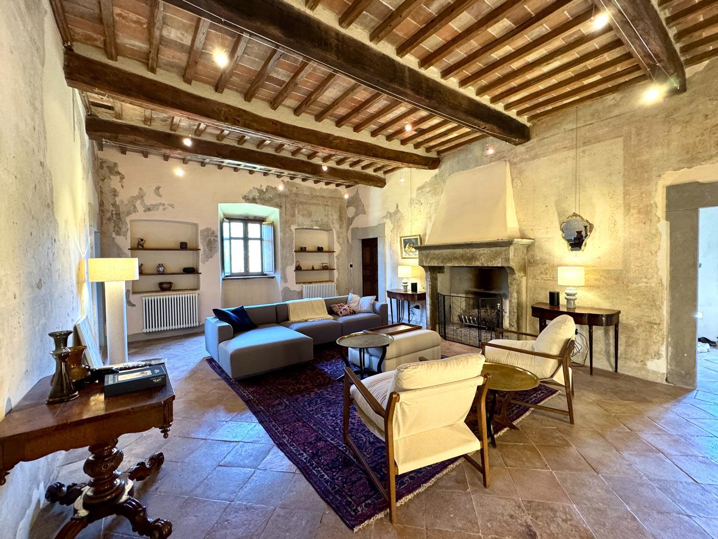 A high-beamed living room with a stone fireplace, modular sofa, and ivory armchairs around a Persian rug