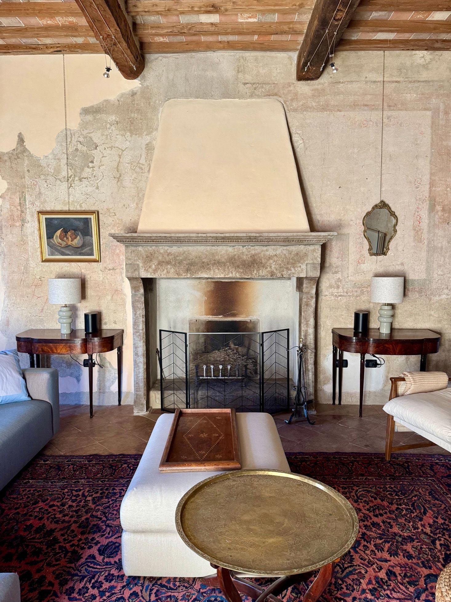A restored living room with a stone fireplace beneath a tall plaster hood, ceramic lamps on dark side tables flanking the hearth, layered Persian rugs underfoot