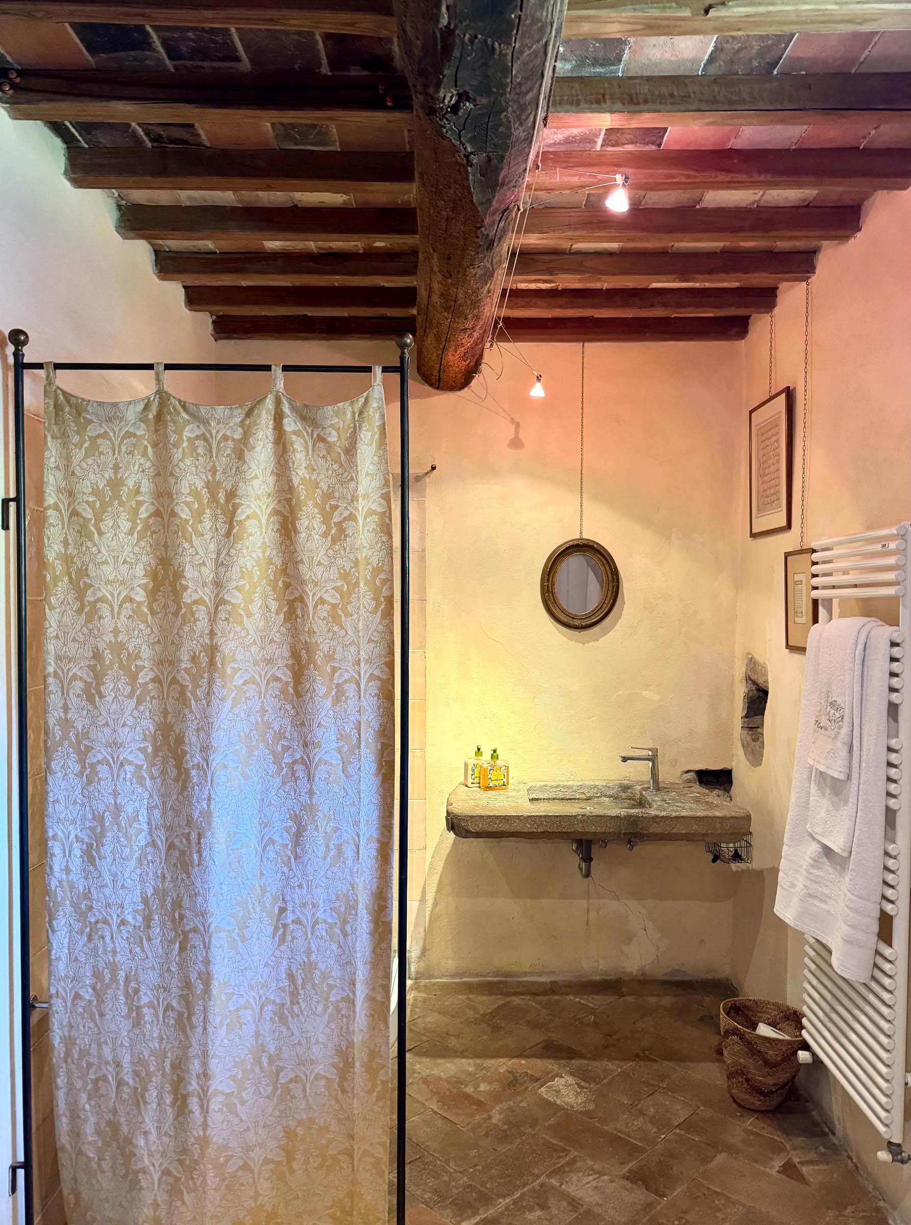 Bathroom with a damask-patterned shower curtain, a long carved stone trough sink and oval gilt mirror beneath a beamed ceiling