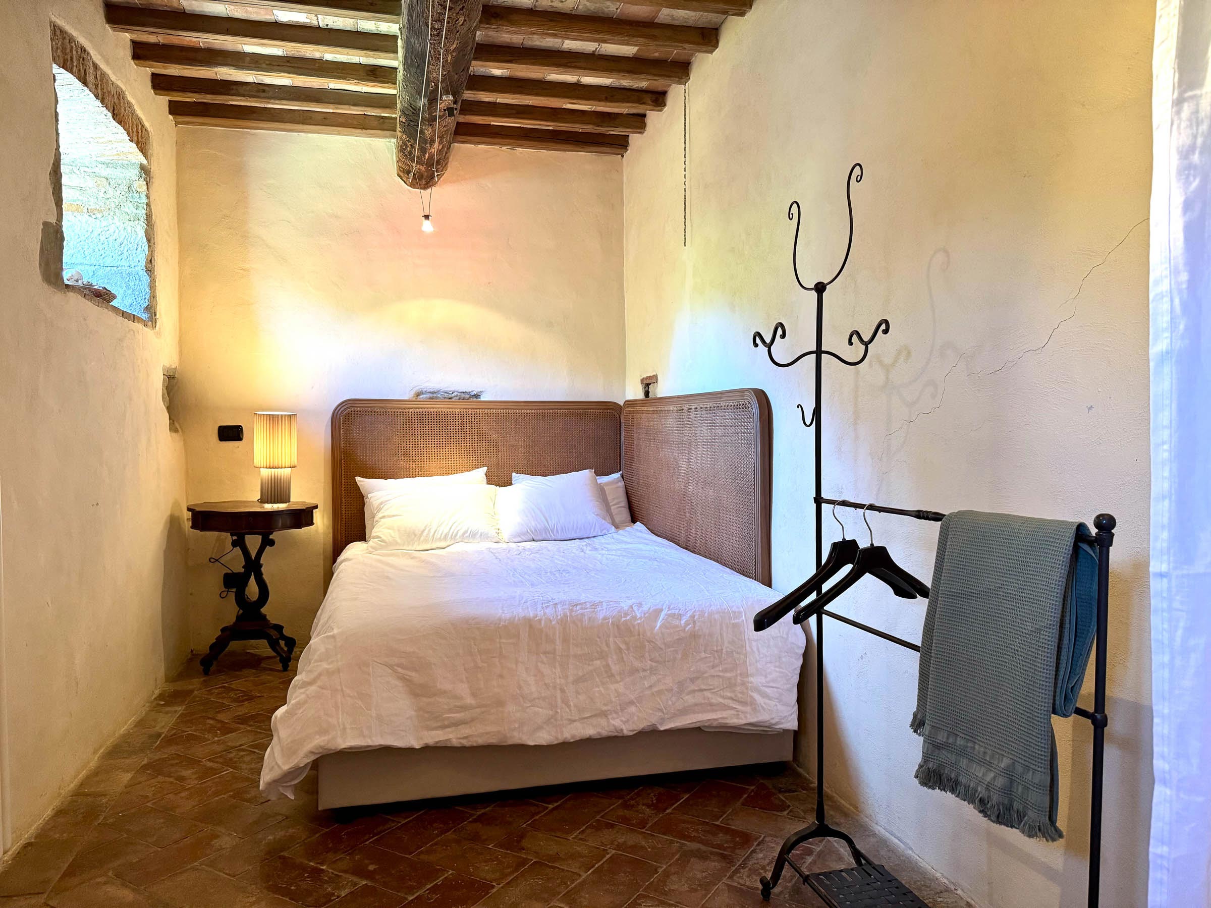Double bed with a wicker headboard against pale plaster walls, beamed ceiling, terracotta floor, an iron coat-stand to one side
