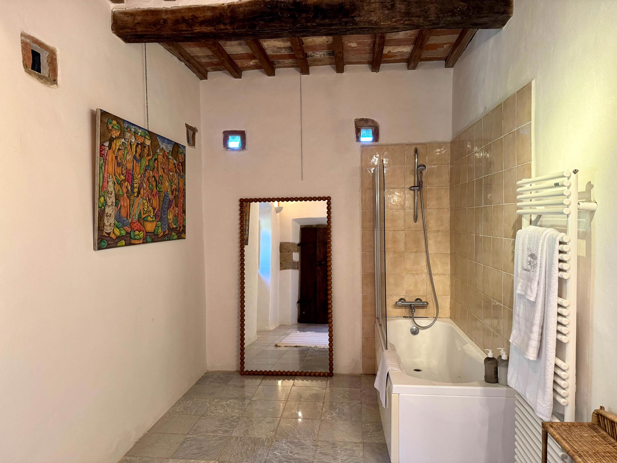 Bathroom with a colourful figurative painting on the wall, a tall framed mirror, and a tiled tub beneath a beamed ceiling