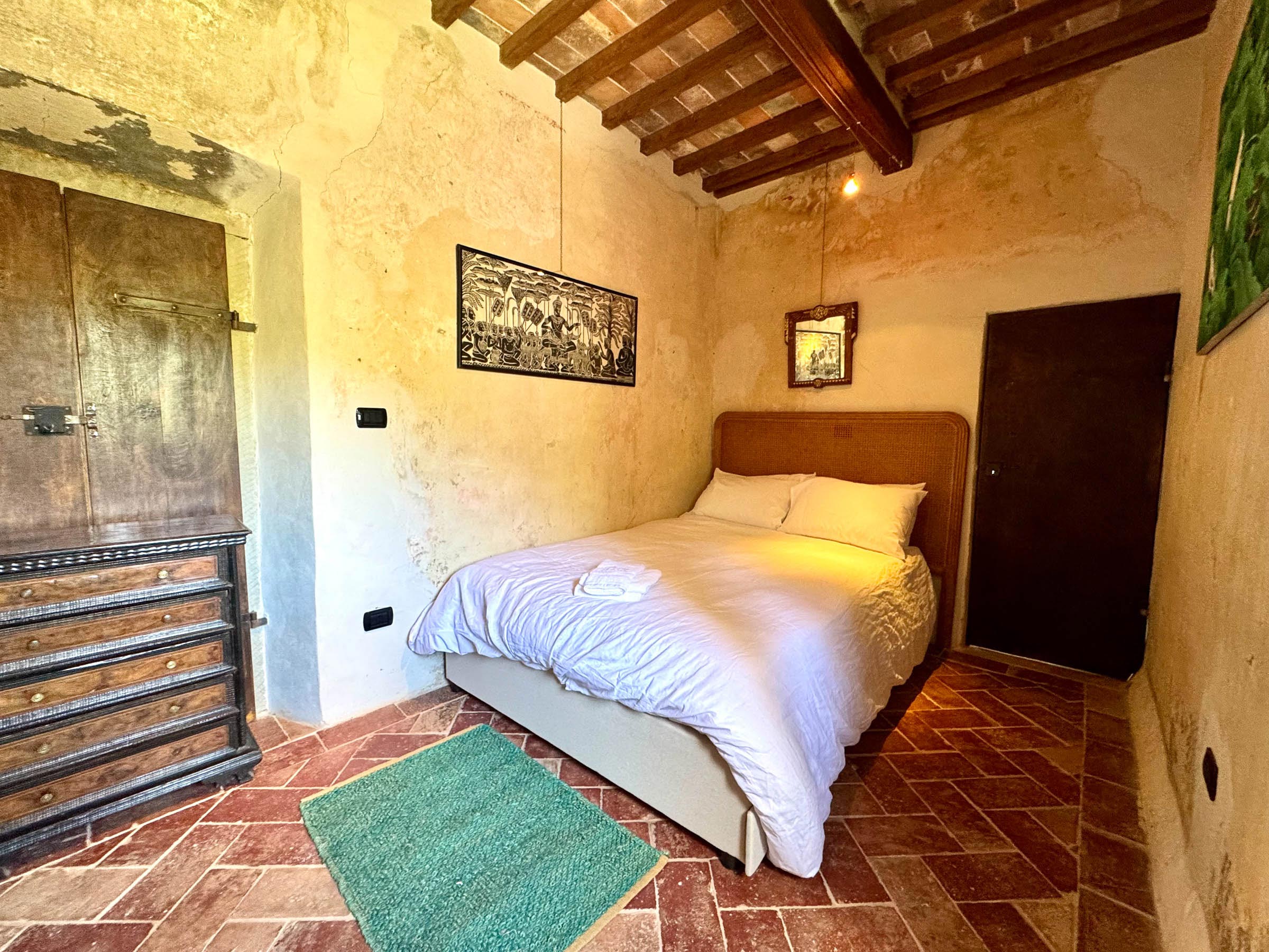 Double bed with white linens beside a tall antique dark-wood chest of drawers, beamed ceiling and terracotta floor