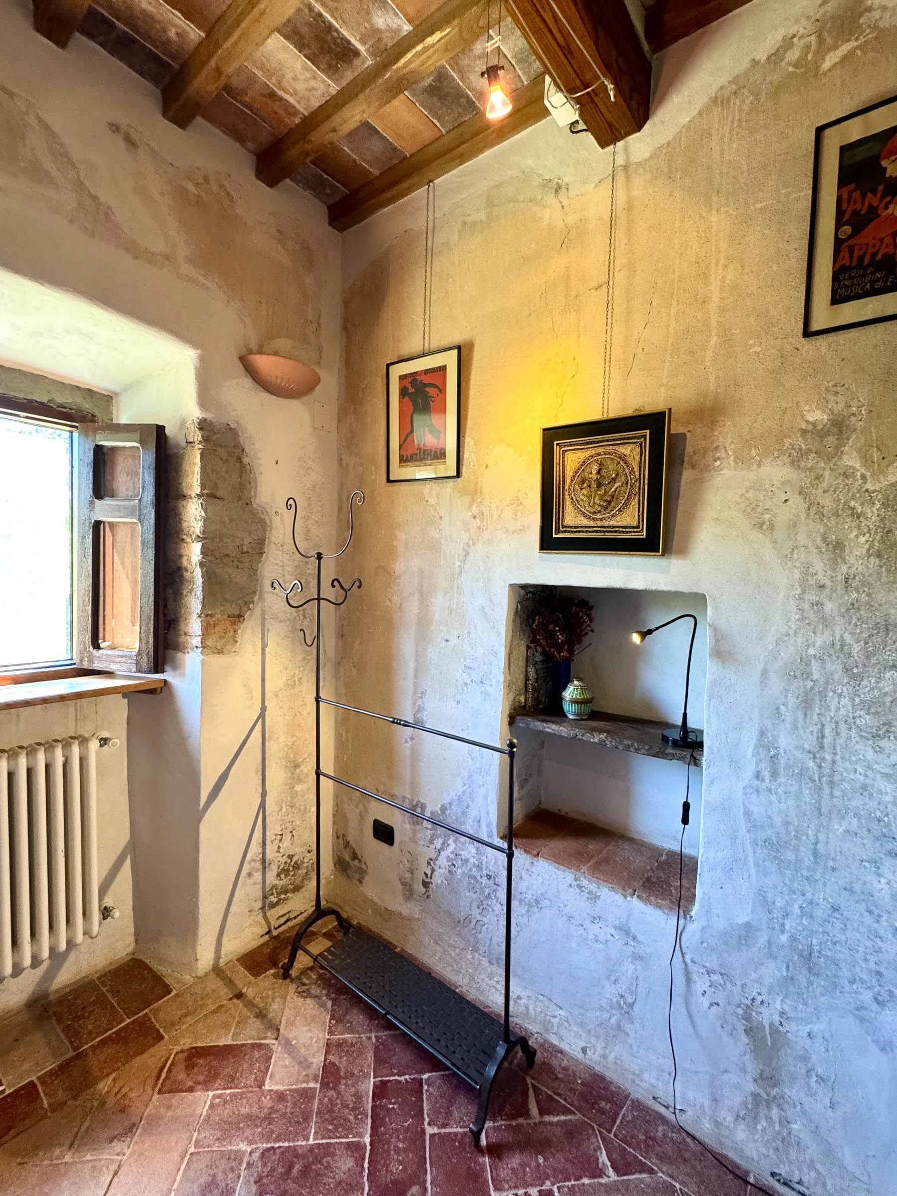 Bedroom corner with a deep stone-framed niche holding a small lamp, framed artworks on plaster walls, iron coat-stand and shuttered window