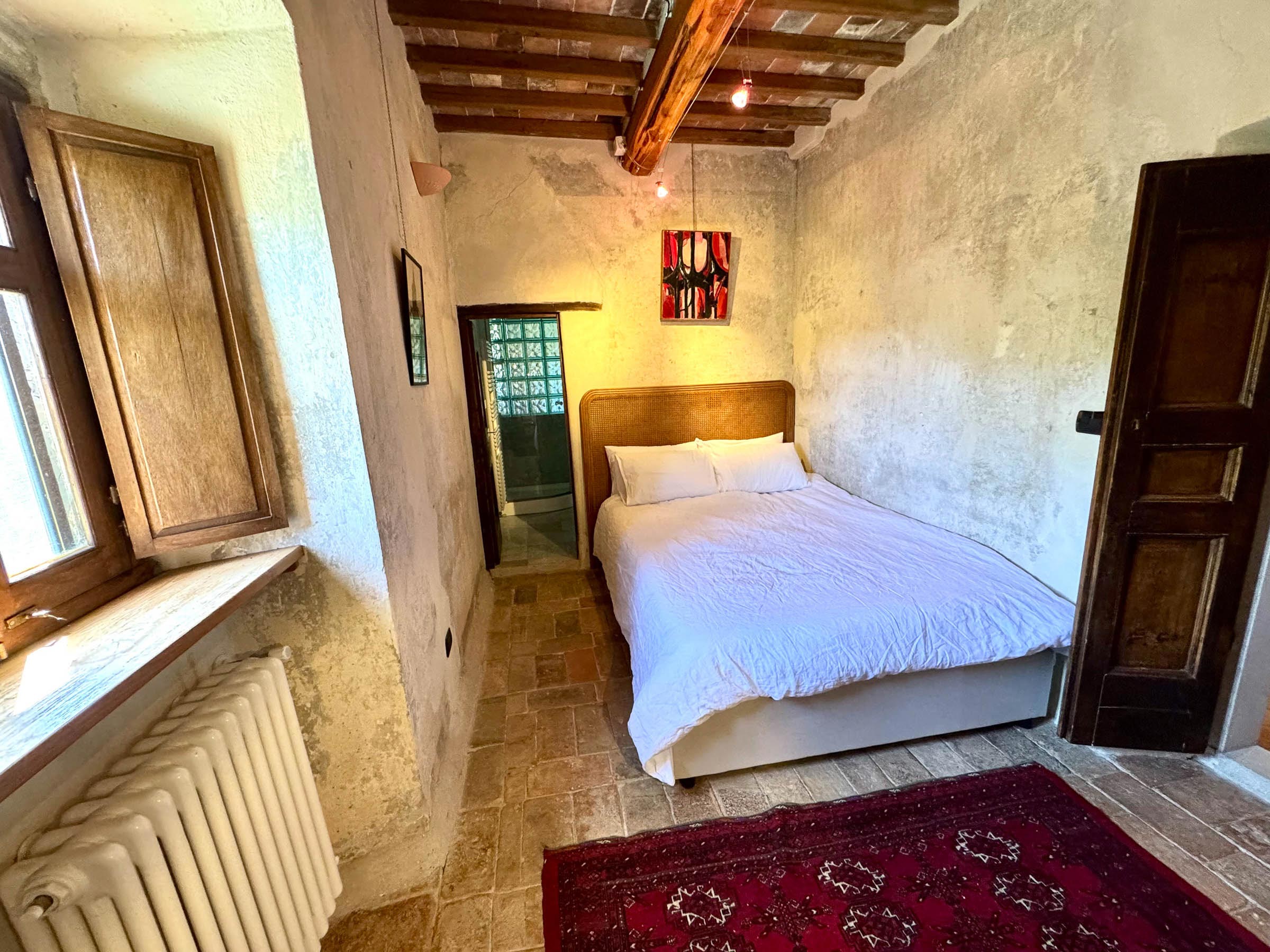 Double bed with white linens beside a deep window recess and a dark wooden door, red Persian rug at the foot, beamed ceiling