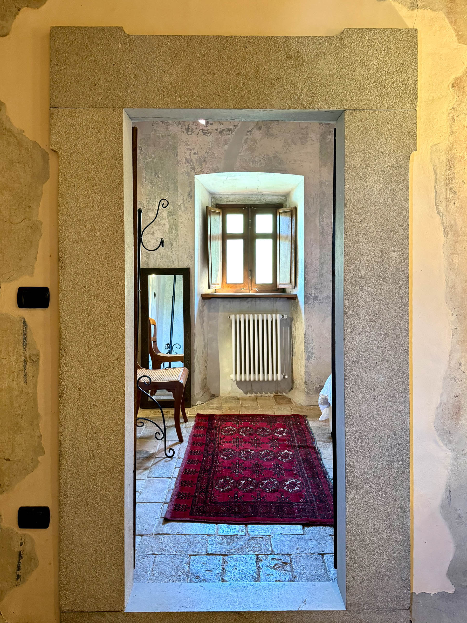 View through a carved stone doorway into a small room with a shuttered window, radiator, and red Persian rug on the stone floor