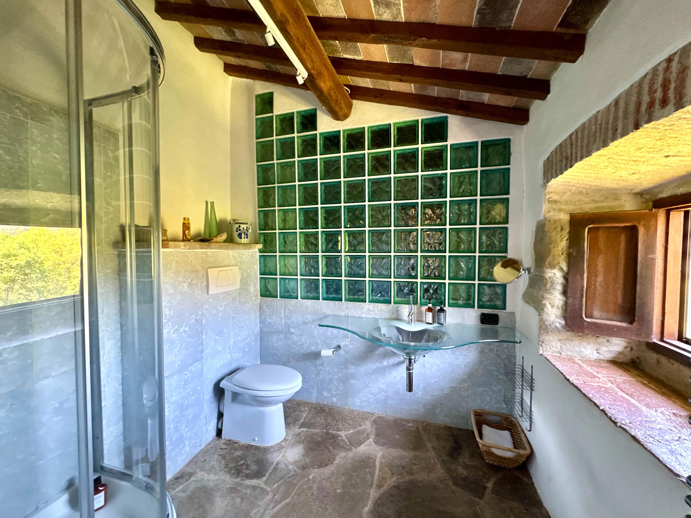 Bathroom with a green glass-block wall, sculptural glass vessel sink on a wooden shelf, rounded glass shower and a deep stone-framed window