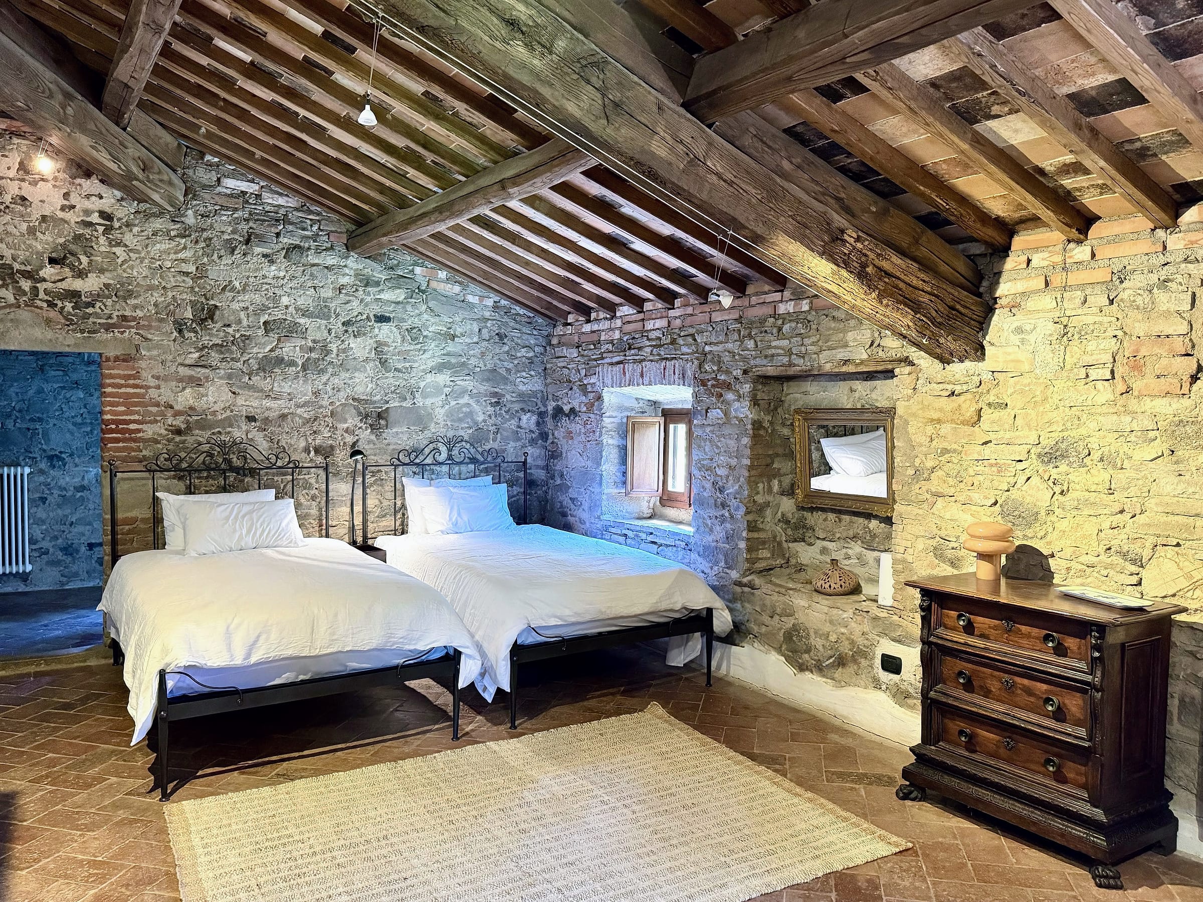 Two wrought-iron small-double beds with white linen against a stone wall, beneath a sloping beamed attic ceiling, an antique chest of drawers alongside