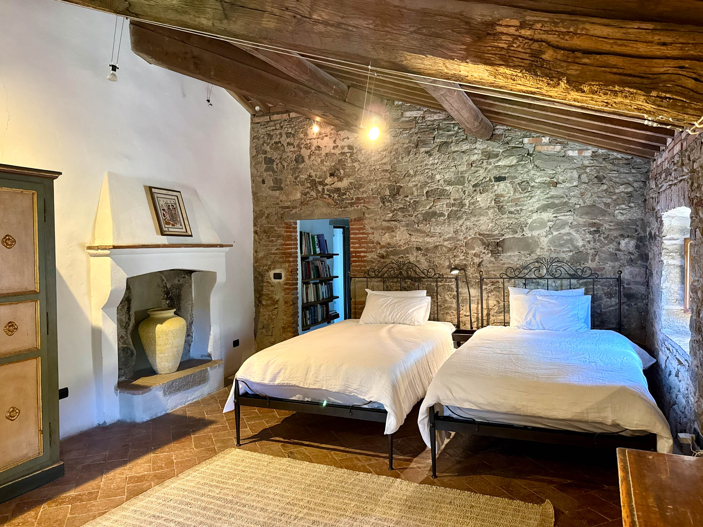Two single beds with iron headboards against a bare stone wall, painted armoire and a small alcove fireplace, beneath a sloping beamed attic ceiling