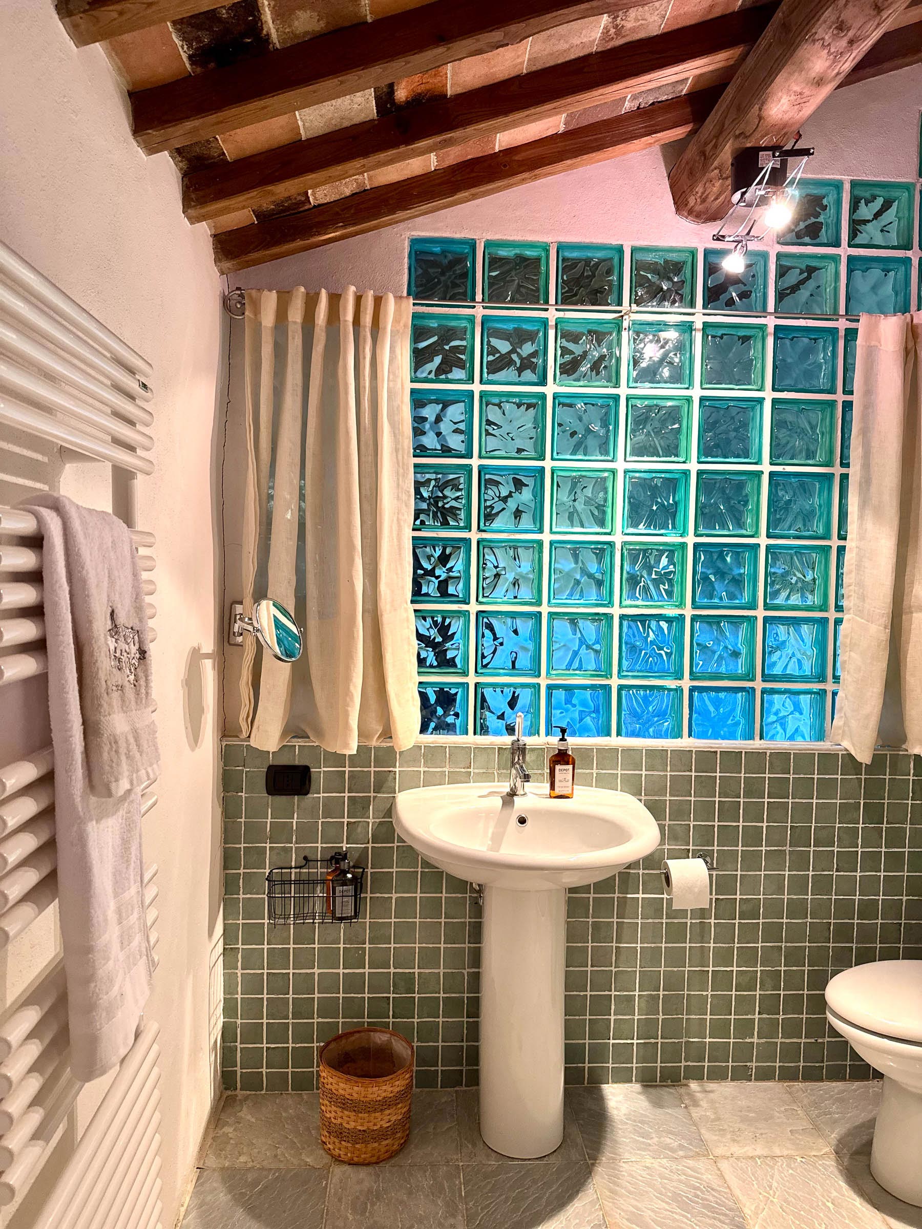 Bathroom with a wall of blue glass blocks behind a curtained window, white pedestal sink and round mirror, green tiled wainscot