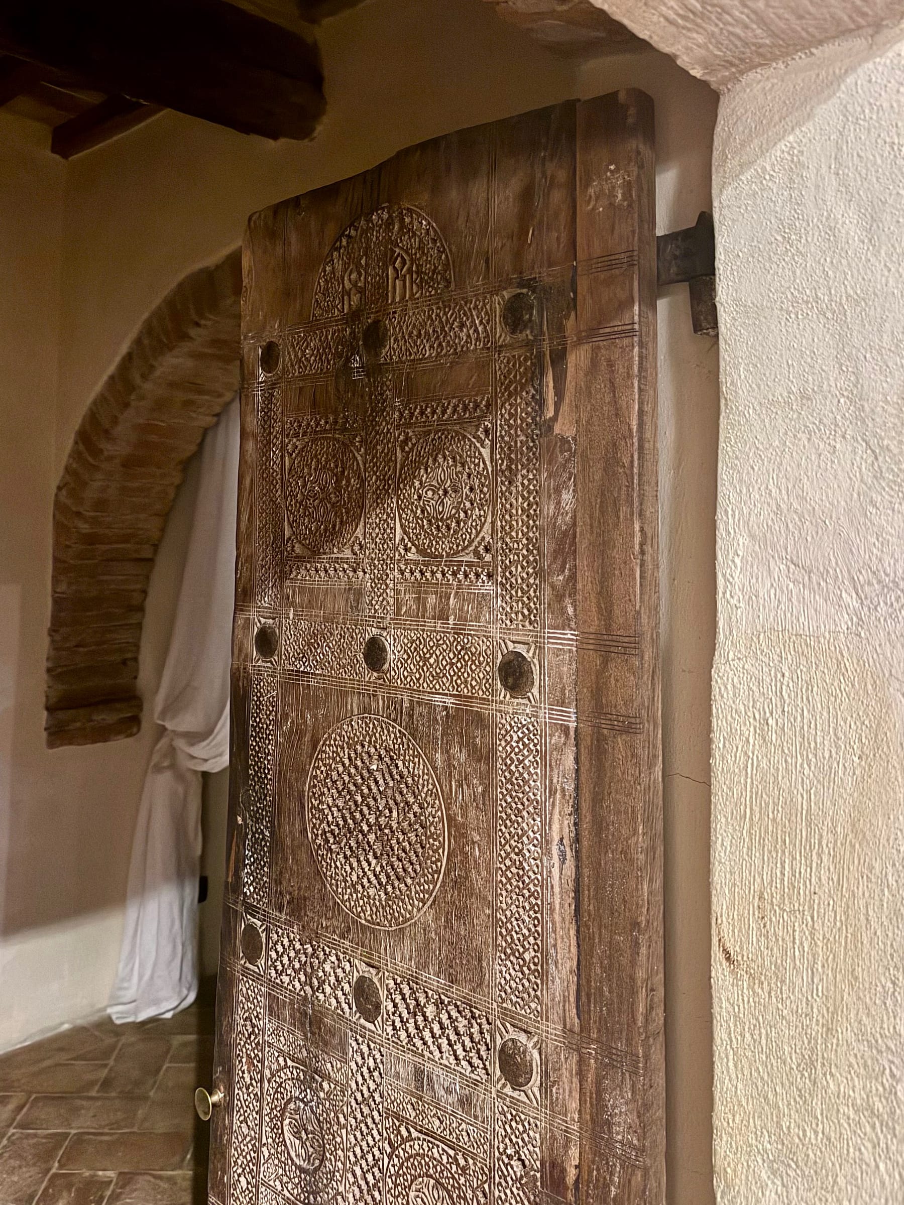 Heavily carved wooden door with geometric and rosette panels, standing open within a brick arch onto a bedroom