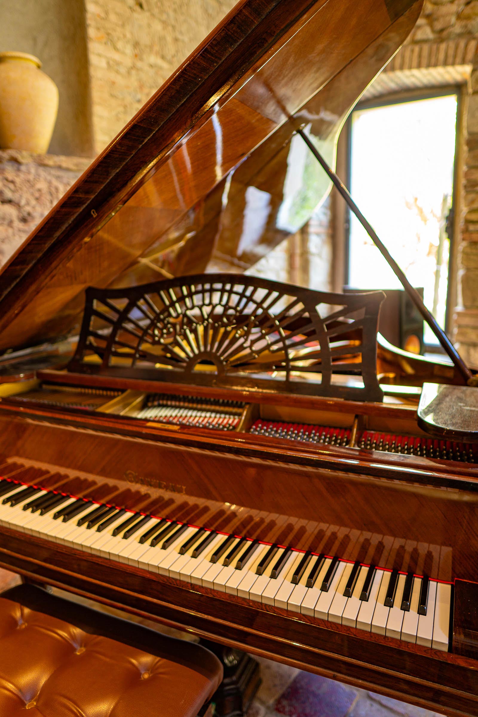 Close-up of an antique grand piano with its lid raised, carved music desk above the keyboard, stone wall and window beyond