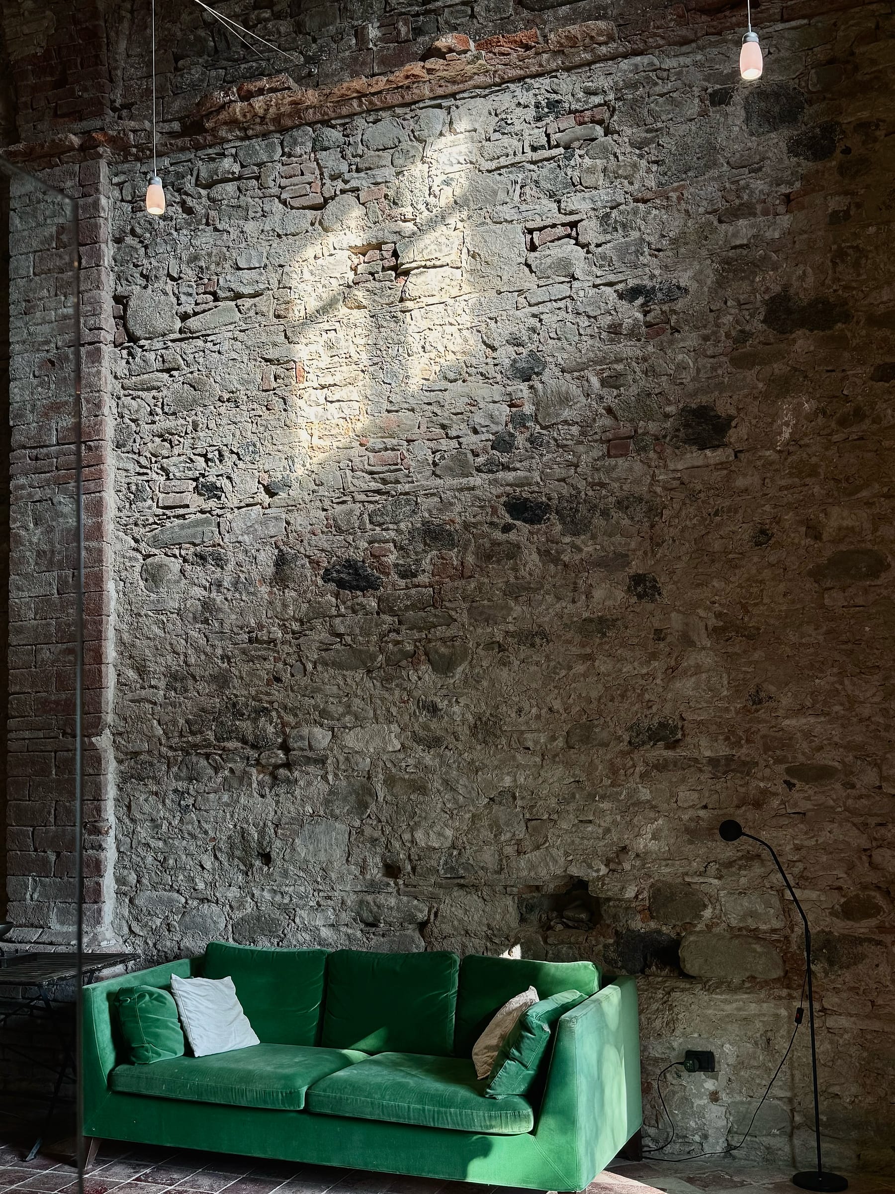 Emerald green sofa against a tall wall of exposed stone and brick, lit by two hanging bulbs