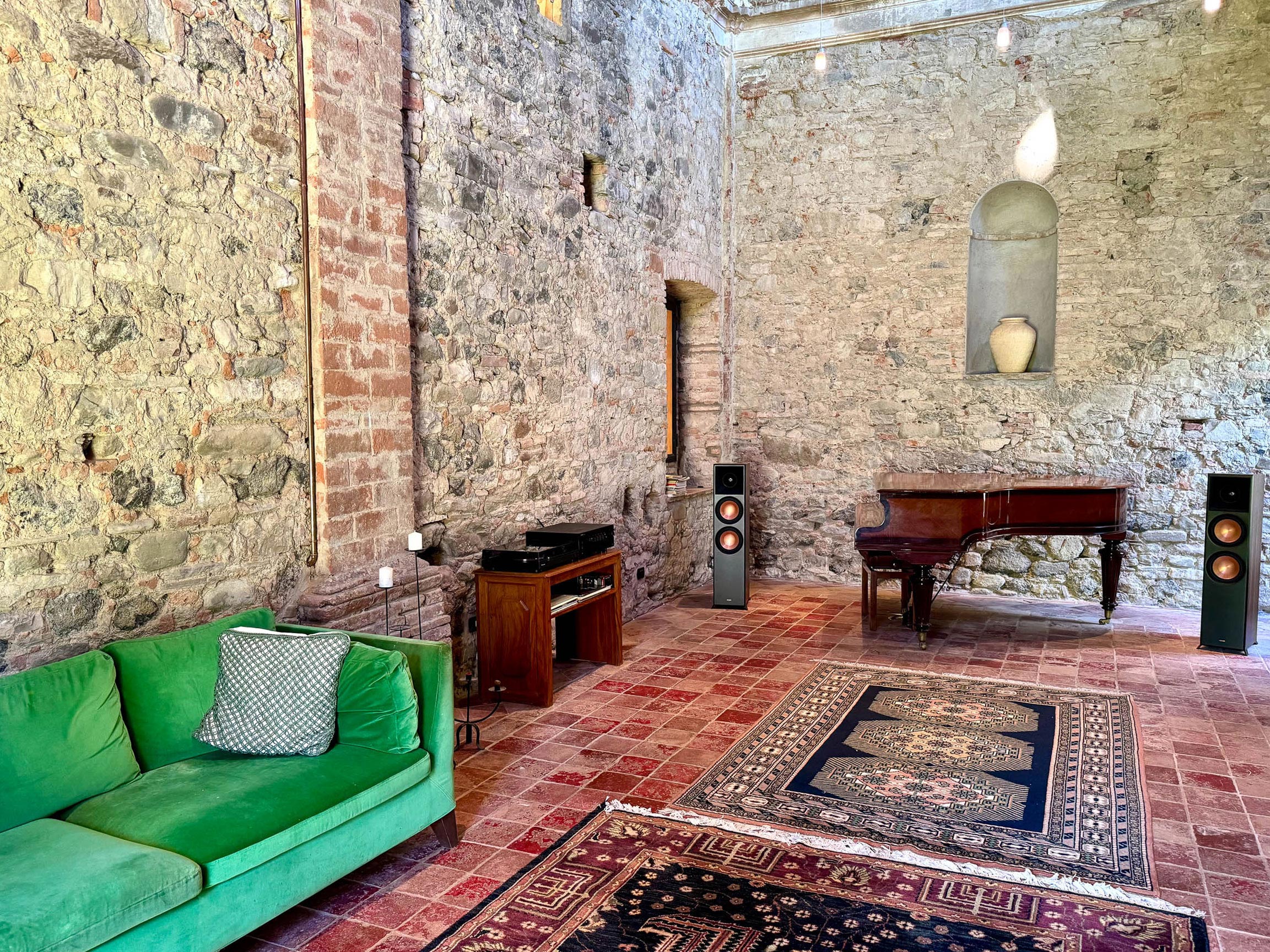 Converted chapel with high stone walls, an emerald-green sofa, turntable cabinet with speakers, and an antique piano on layered Persian rugs