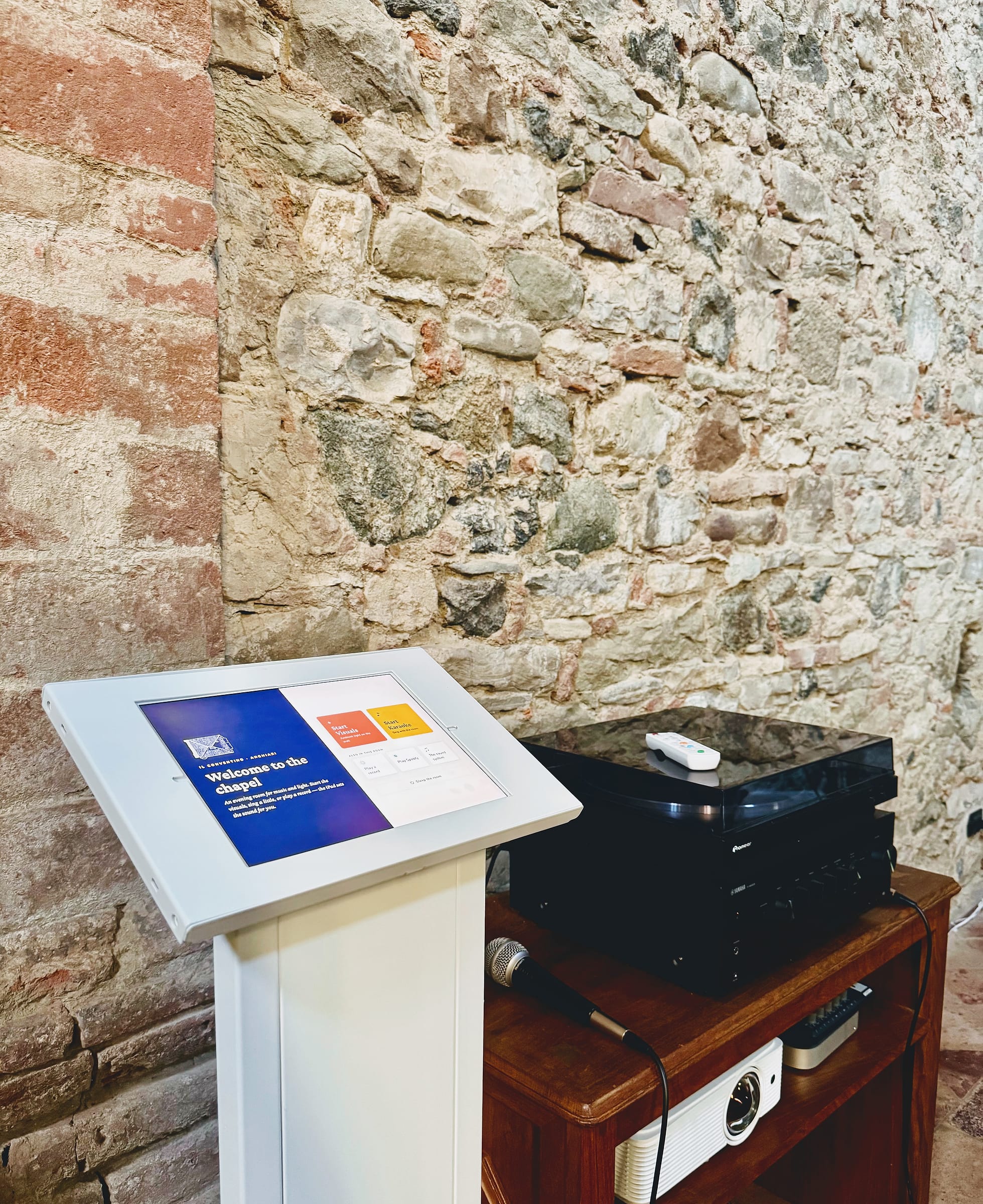 A tablet kiosk on a white stand beside a wooden trolley holding a turntable, an amplifier, a microphone and a projector, against a wall of stone and brick