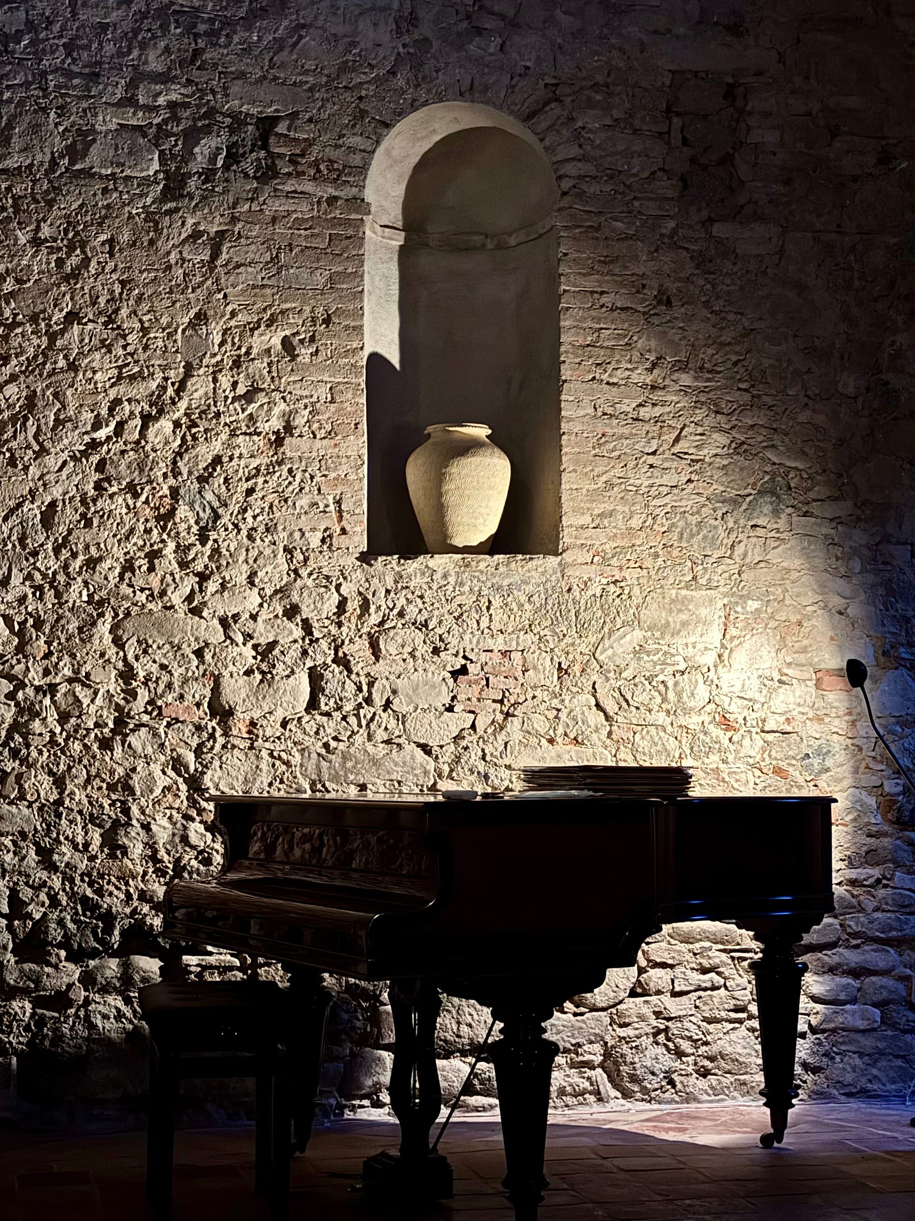 Baby grand piano in low evening light against a rough stone wall, a pale urn lit in a sculpted niche above