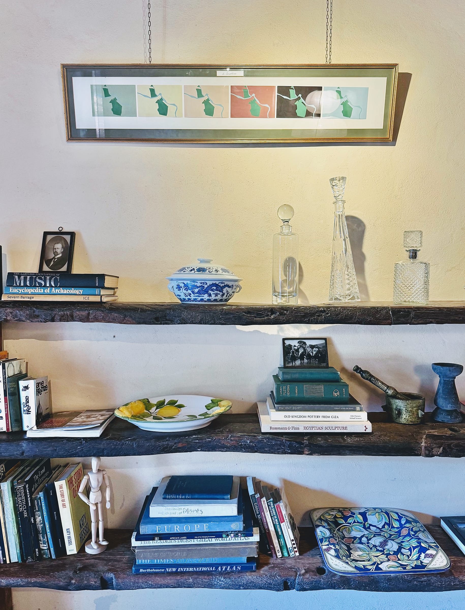 Books, blue-and-white china and glass decanters arranged along three rough timber shelves below a long framed print