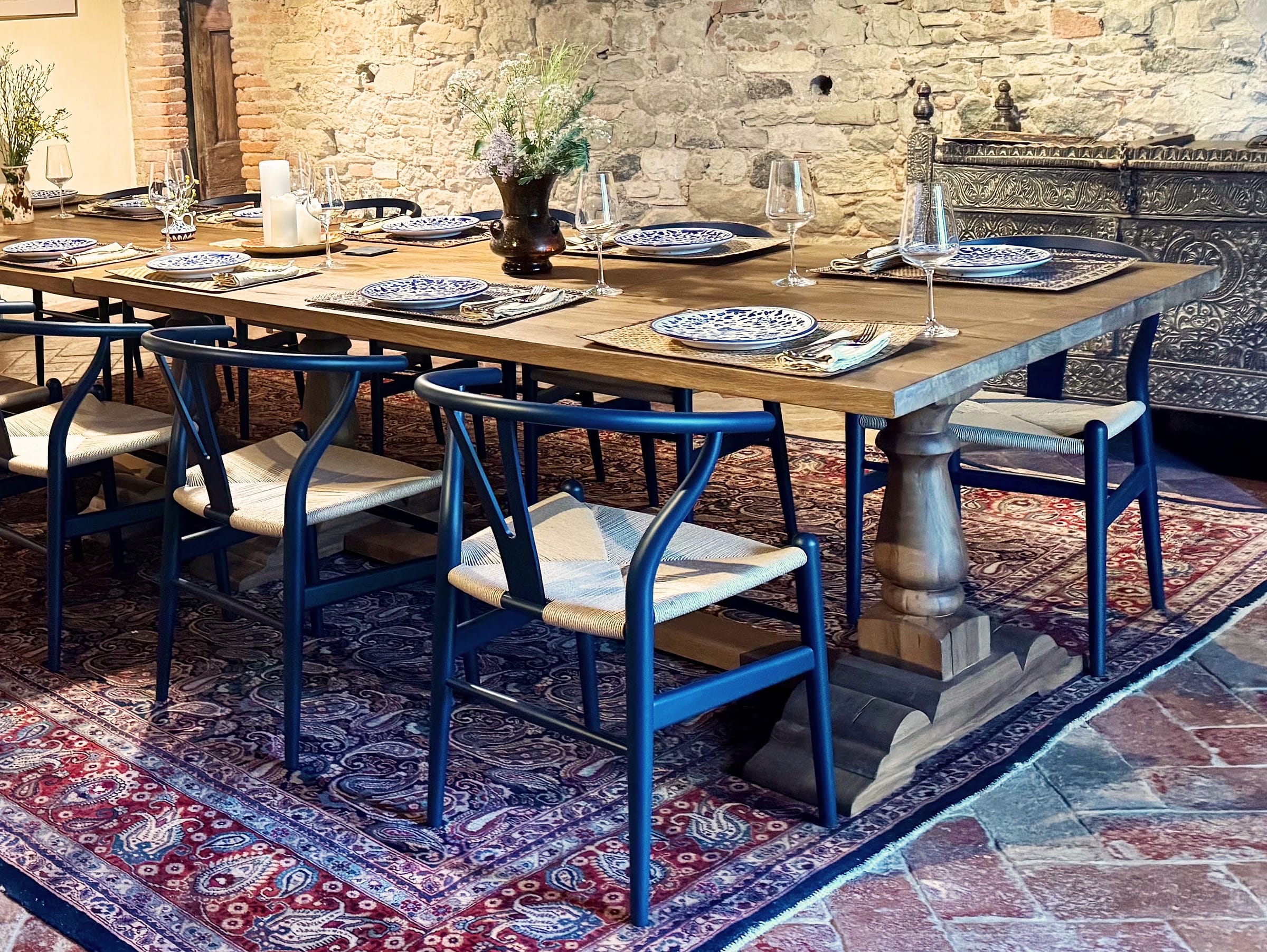 Two long pale wood tables placed end to end, laid with blue-and-white plates and wine glasses, blue wishbone chairs around them, a carved dark chest against the stone wall behind
