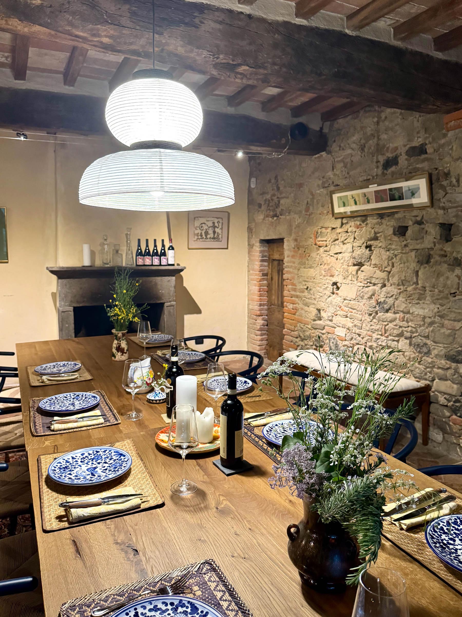 Long wooden dining table set with blue-and-white plates beneath a paper pendant lamp, beamed ceiling and exposed stone wall