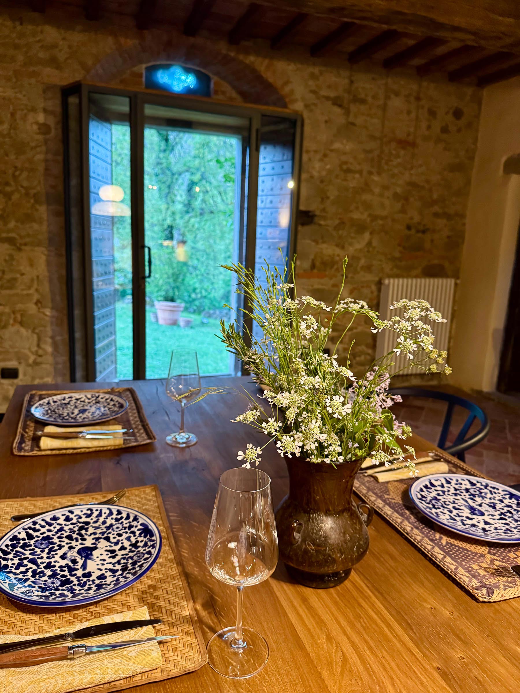 Wooden table set for dinner with blue-and-white patterned plates and a vase of wildflowers, arched glass doors opening to the garden