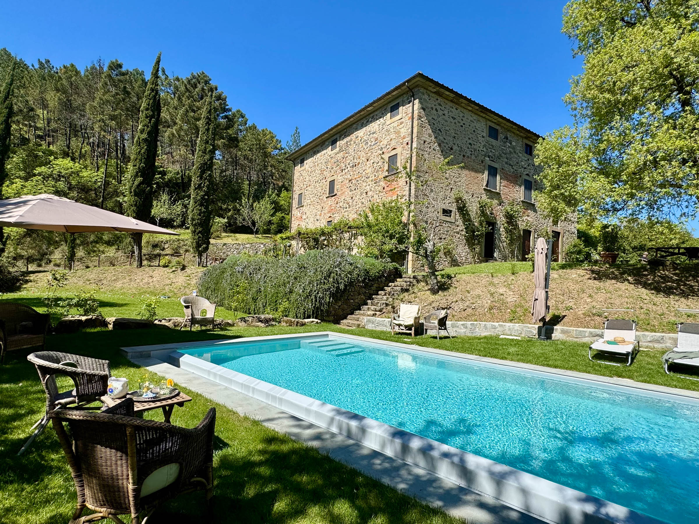 Stone building on a hillside above a swimming pool, sun loungers at the pool edge and wicker chairs in the foreground