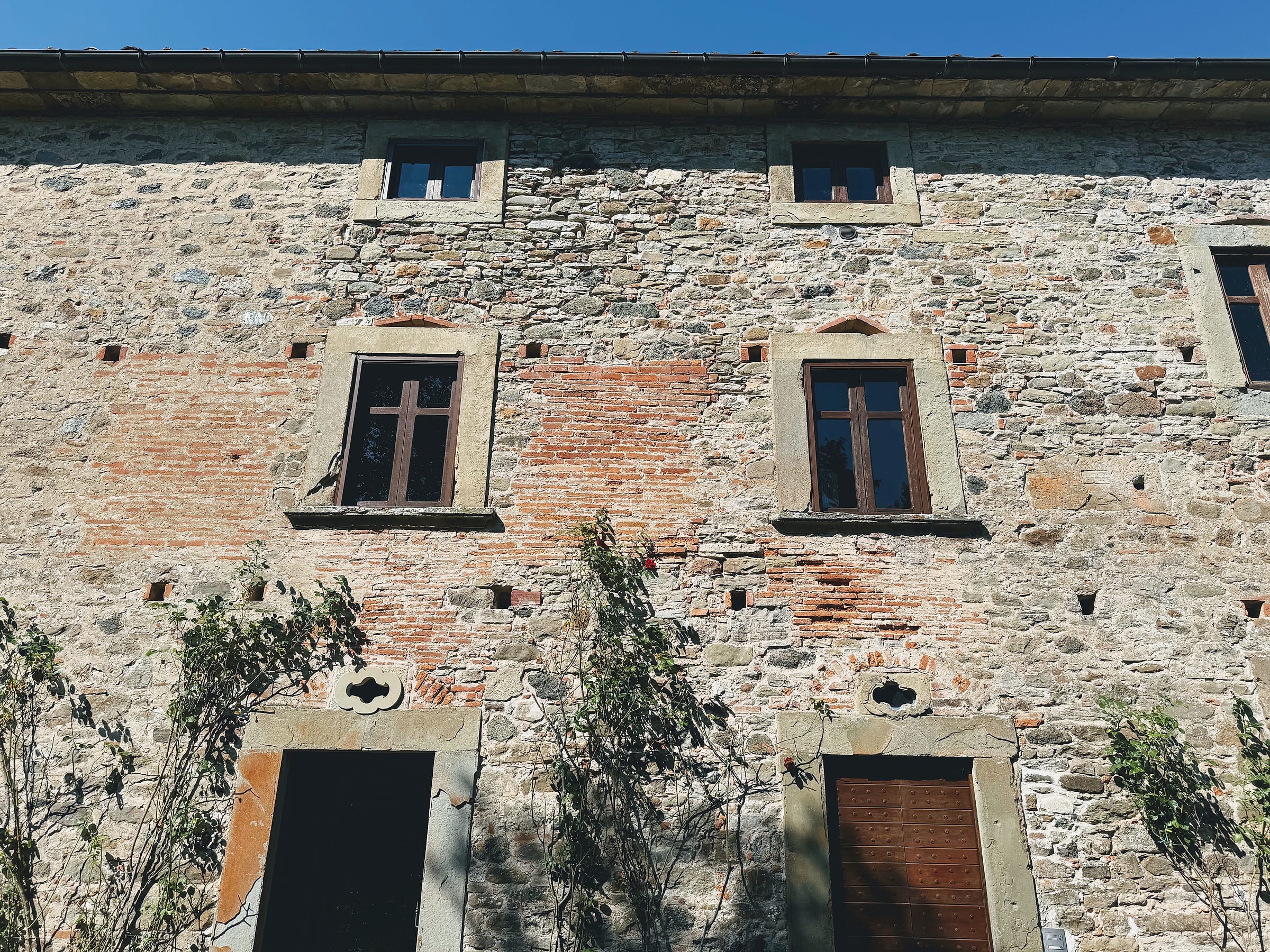 Looking up at a stone and brick facade with two windows in carved stone surrounds, a lobed motif carved above each doorway below, and rows of small square putlog holes across the wall