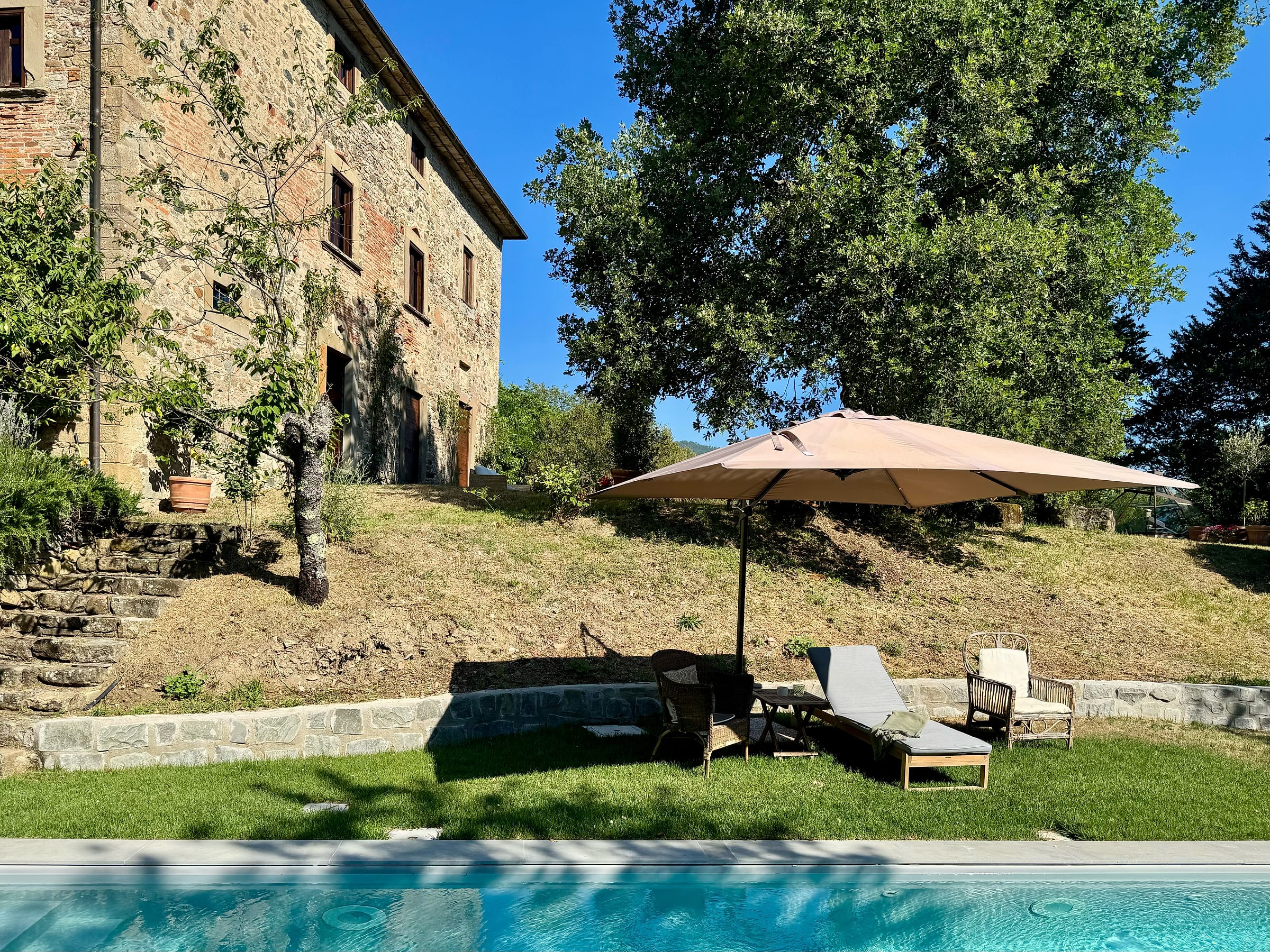 The stone building on the slope above the pool, with a parasol and sun loungers on the lawn between them