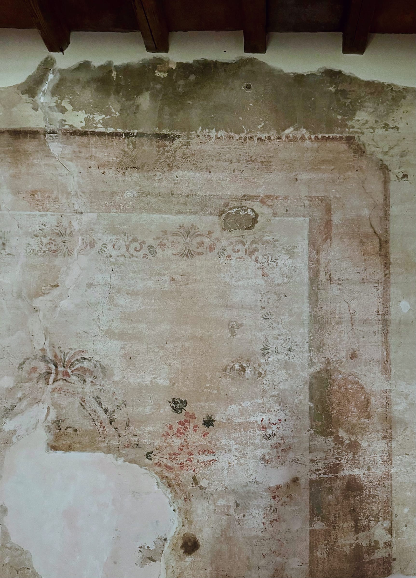 Faded fresco with a scrolled border band, painted frame lines and floral sprigs on an old plaster wall beneath dark beams