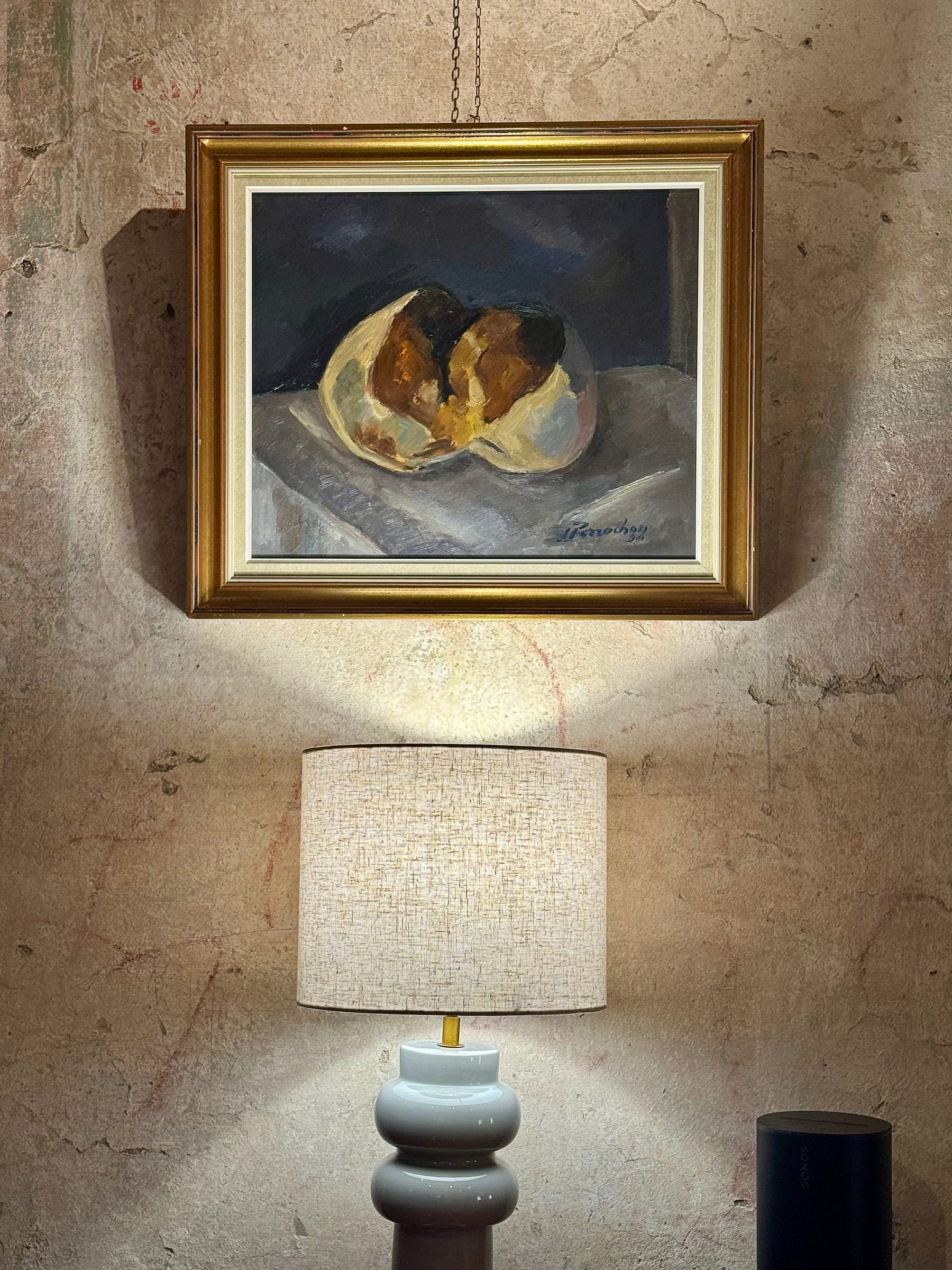 Gilt-framed still life hanging from a chain on a wall of aged plaster, above a table lamp with a pale bulbous base and a wide pale shade