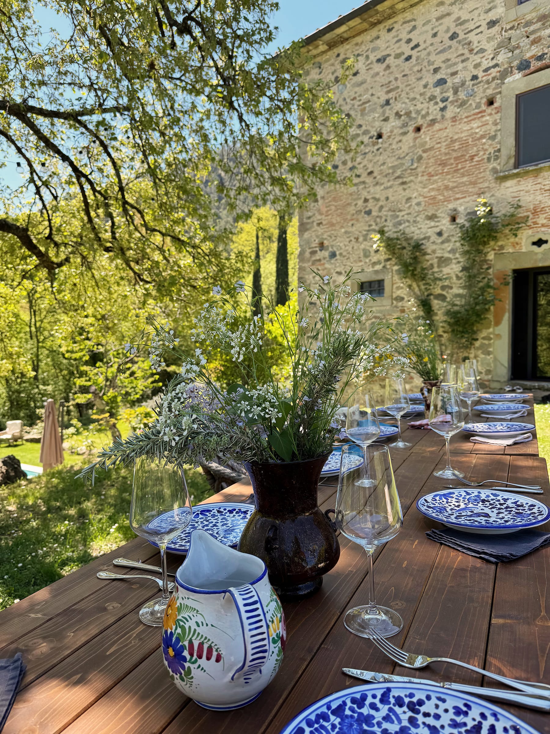 A long outdoor table laid with blue-and-white plates and wine glasses under trees, the stone building rising behind