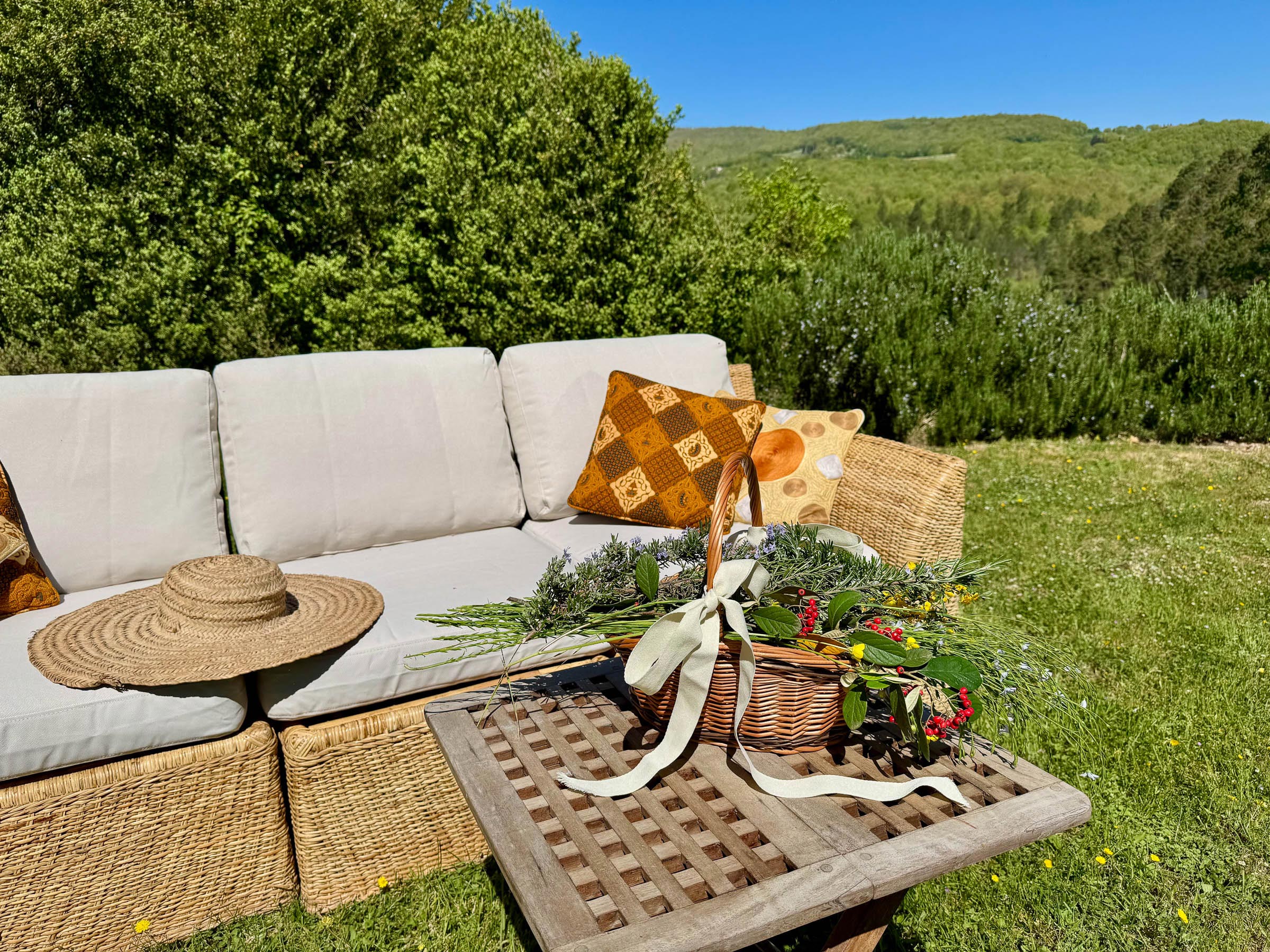 Wicker outdoor sofa with a straw sun hat resting on the cushions, a basket of foraged flowers on a slatted wooden table, hills rolling away in the distance