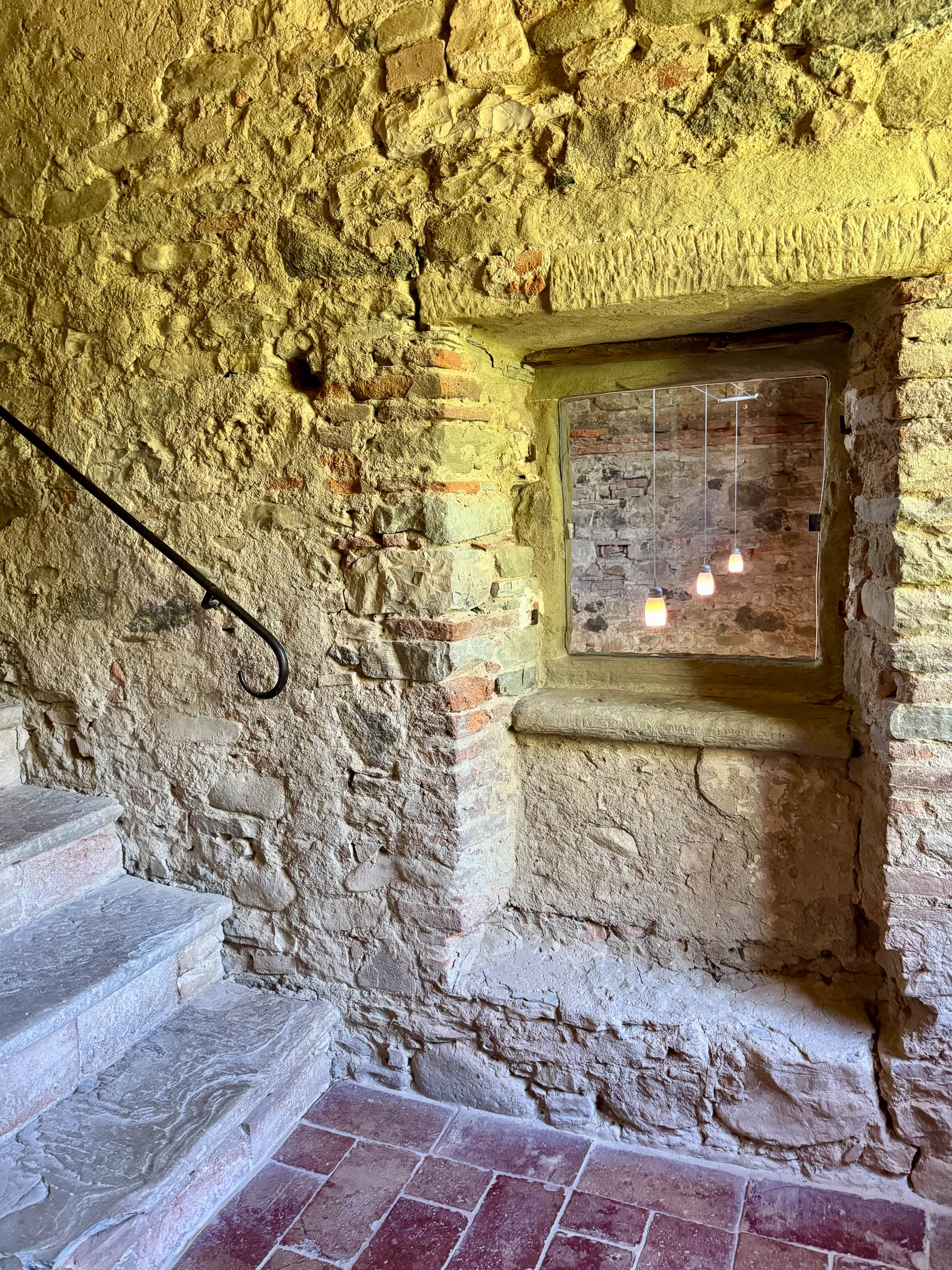 Stone-and-brick interior staircase with an iron handrail, a deep niche looking into the chapel, and a terracotta tile floor