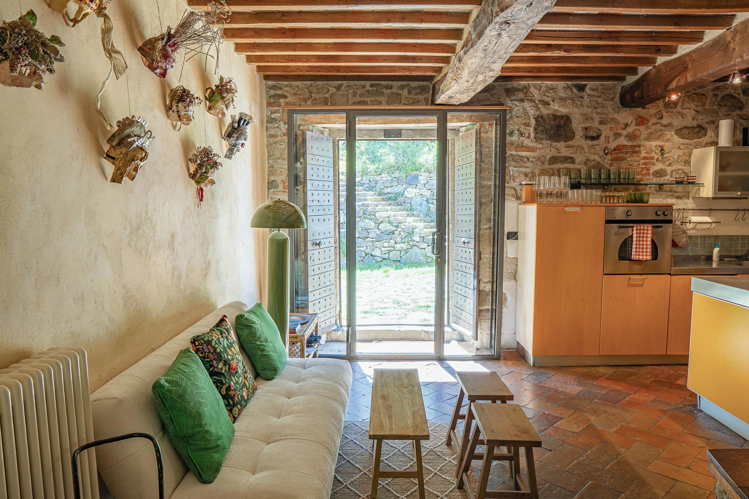 Cream sofa beneath dried botanical Shakespearean masks on the wall, glass-paneled door open to a stone-walled garden, modern yellow kitchen units to the right