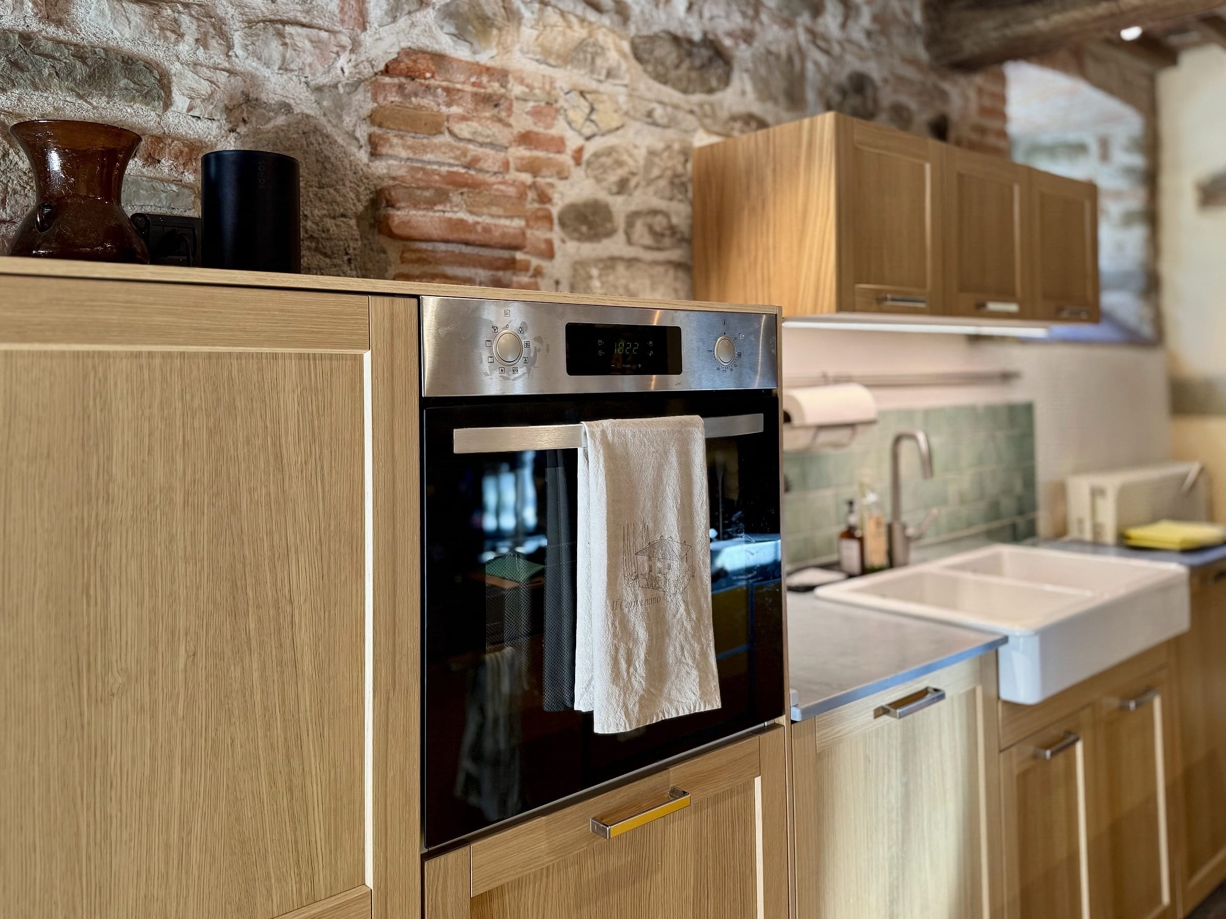 Pale wood kitchen units with a built-in oven and a double white ceramic sink against a green tiled splashback, exposed brick and stone wall behind