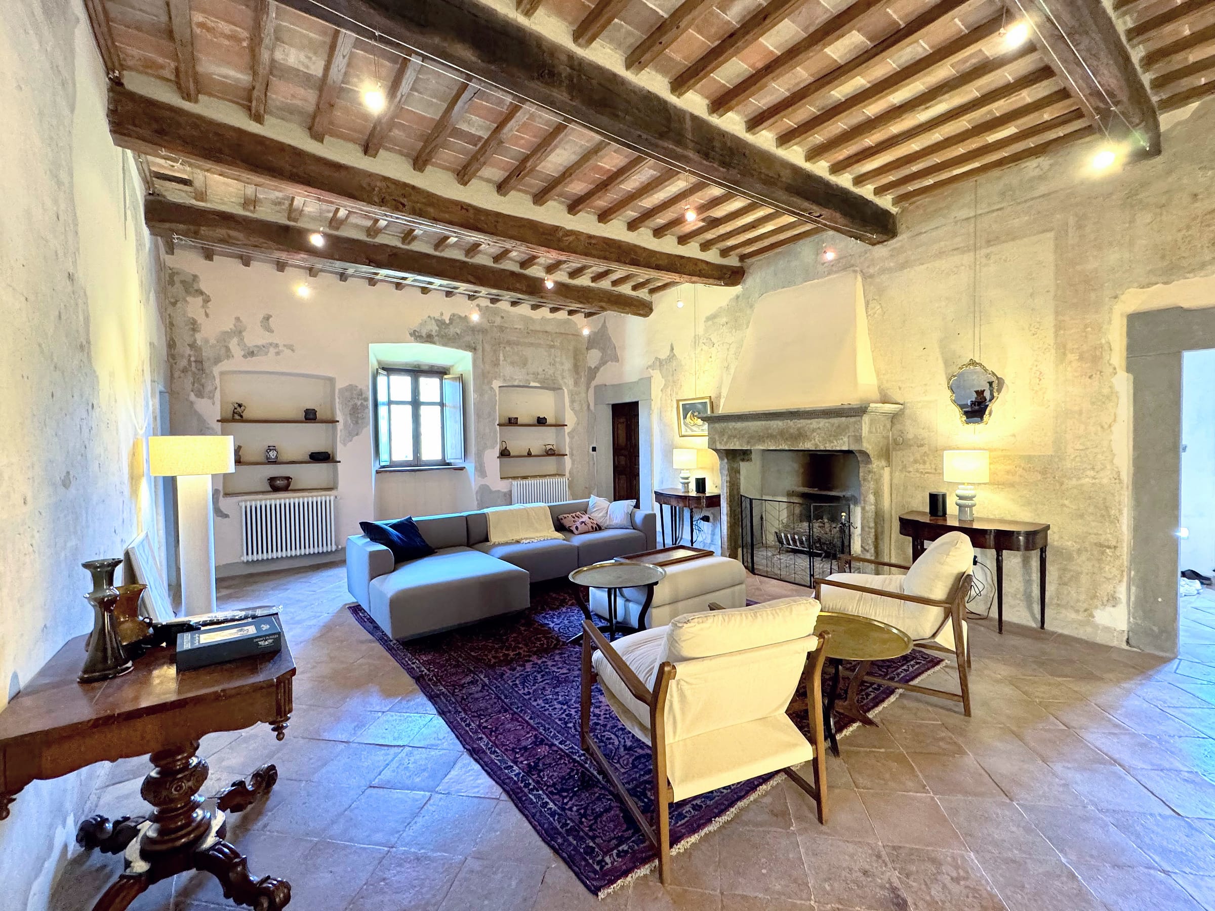 Living room with a stone fireplace under a white plastered hood, a grey sofa, a pale ottoman and two wood-framed armchairs on a patterned rug, beneath a high beamed and tiled ceiling