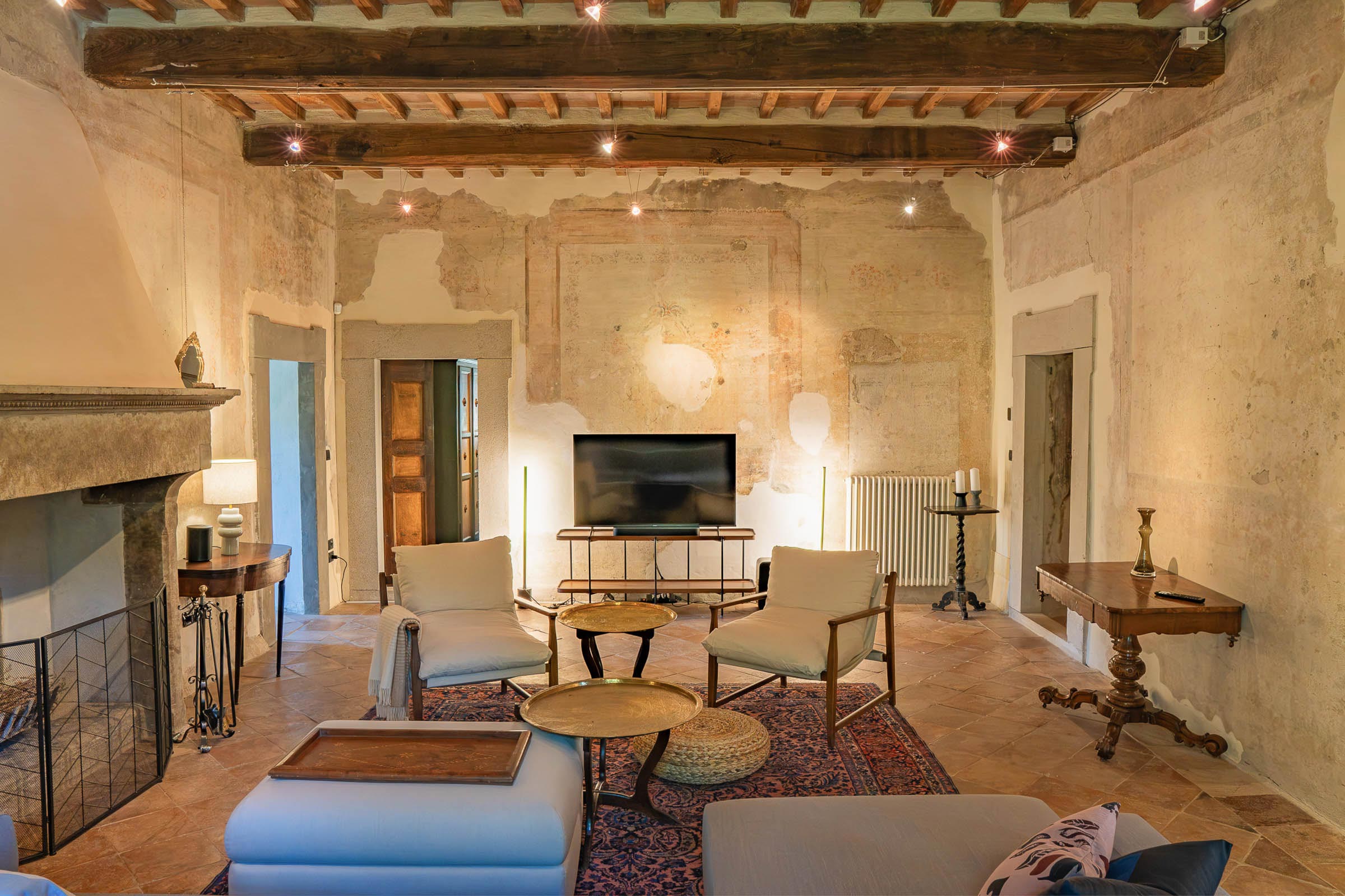 Tall living room with a stone fireplace, two armchairs and a low round table on a Persian rug, beneath a high beamed ceiling