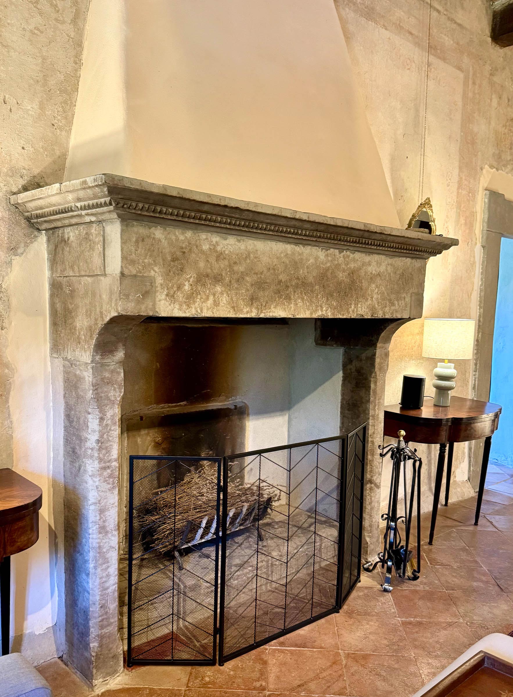 Carved stone fireplace with a conical plaster hood, iron fire screen and tools, kindling stacked inside the hearth