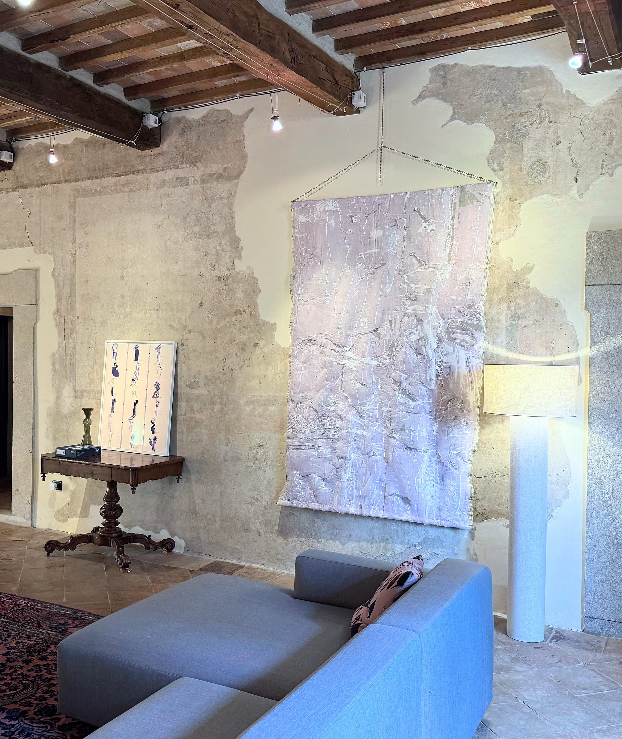 Large pale woven hanging suspended on a cord against a wall of aged, patched plaster, a framed print propped on a carved wooden console to one side, a grey modular sofa in the foreground