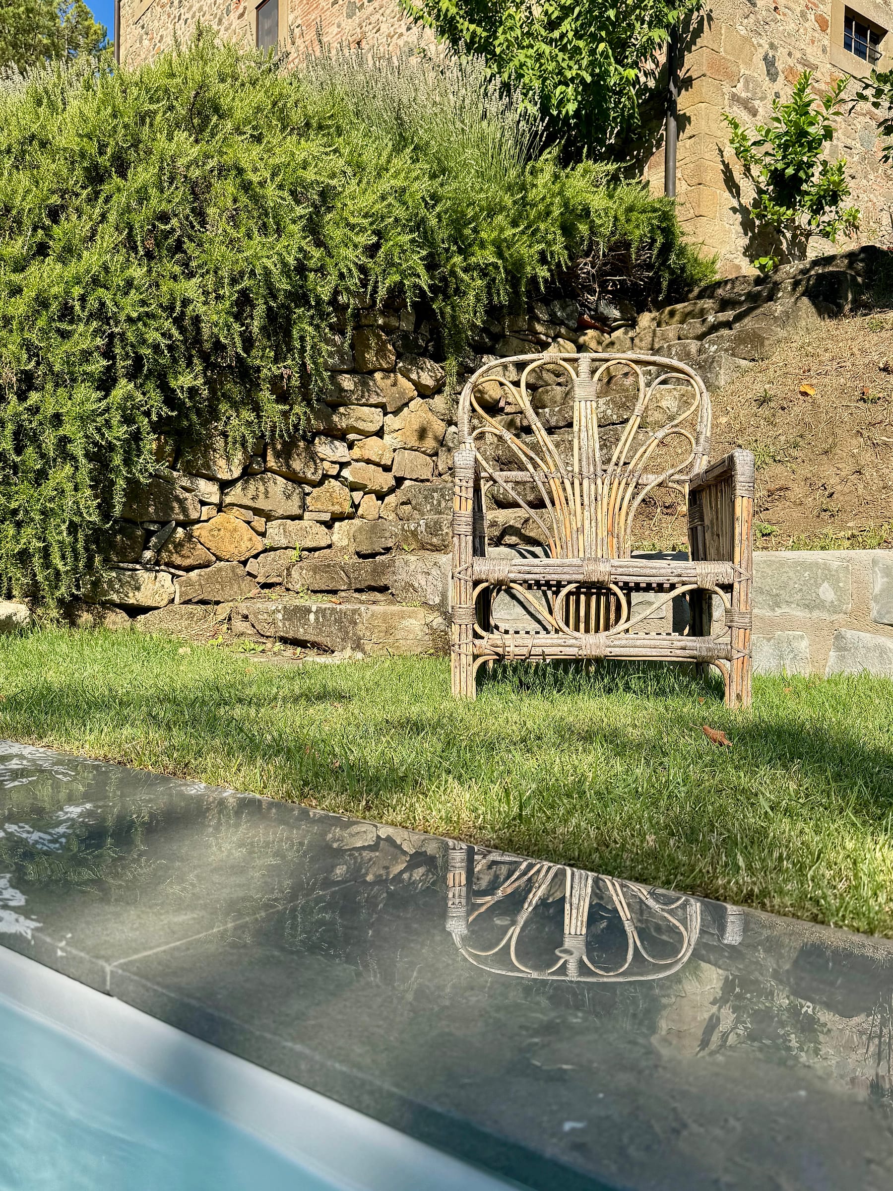 A weathered wicker armchair on the grass between the pool and a dry-stone wall, the stone house rising behind