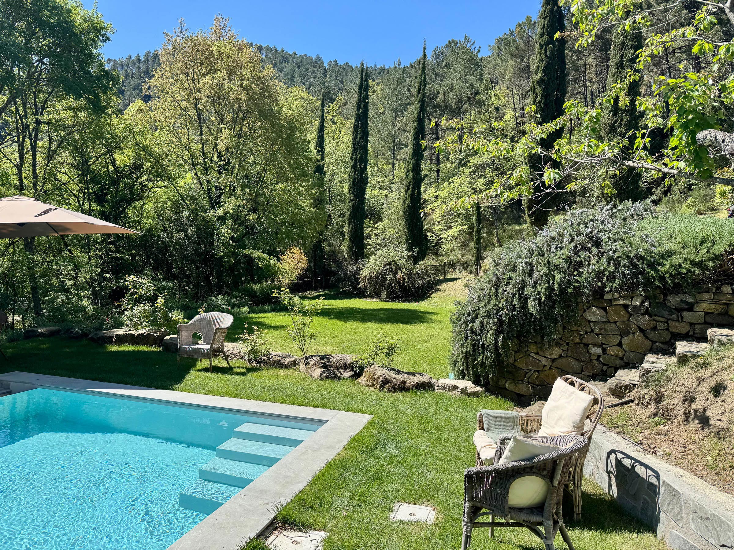 Swimming pool with wicker chairs on the lawn, cypresses and a dry-stone wall beyond