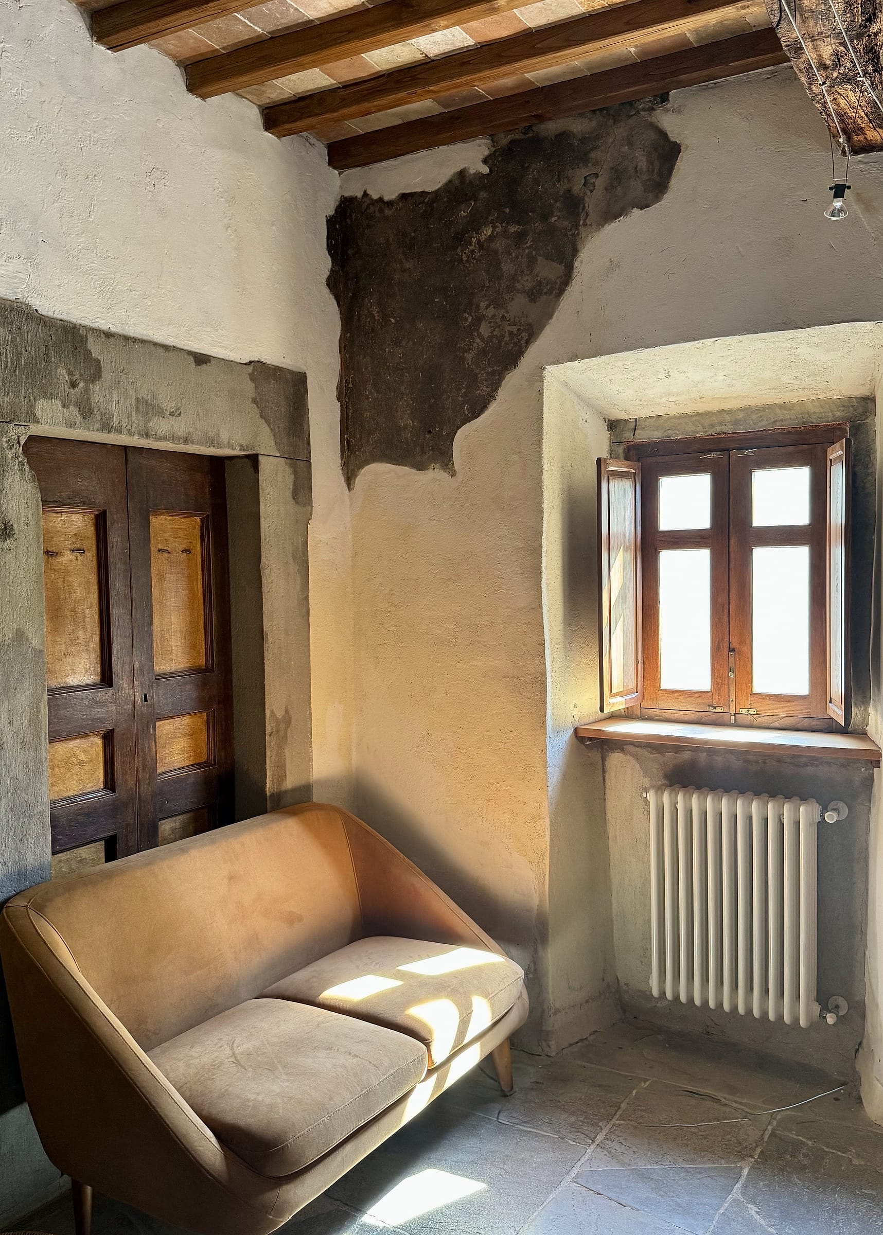 Tan two-seater sofa in a sunlit corner, wooden panelled doors in a stone-framed opening to one side, and a casement window standing open above a radiator in a deep wall recess