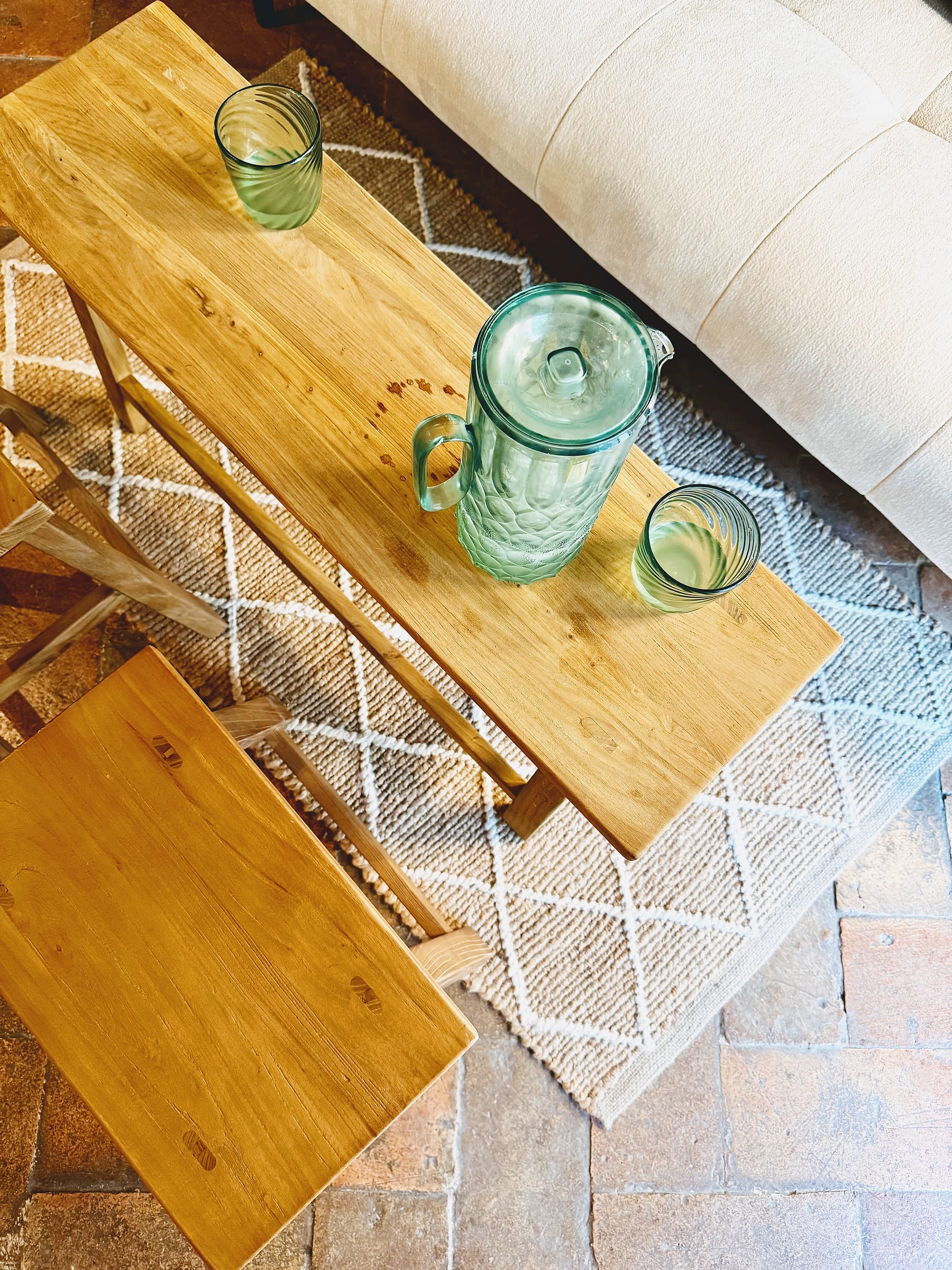 Looking down on two low wooden tables holding a green glass jug and two tumblers, on a patterned rug over a terracotta floor beside a cream sofa