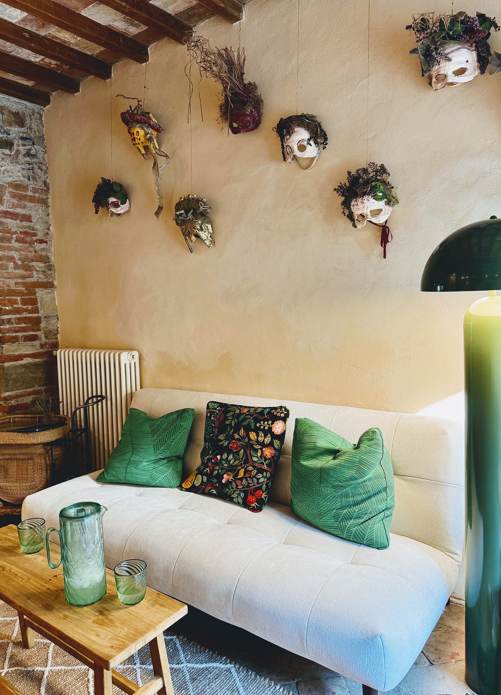 Woven masks hung across a pale plaster wall above a white sofa with green cushions, a green floor lamp to one side