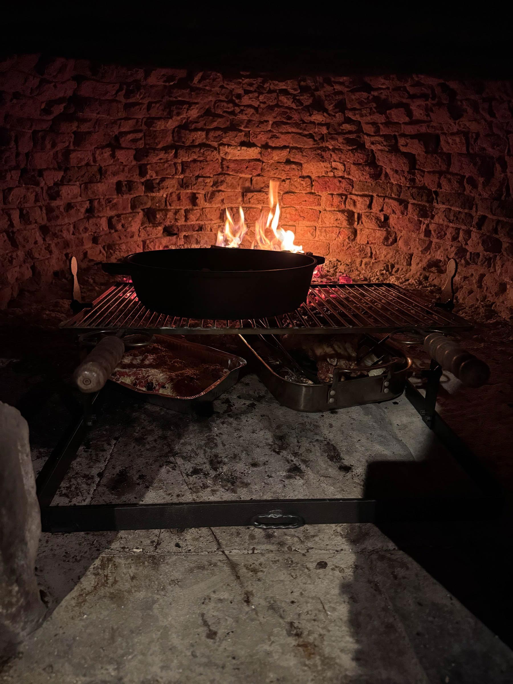 Cast-iron pot on a grate inside the wood-fired oven, flames blazing within the brick interior