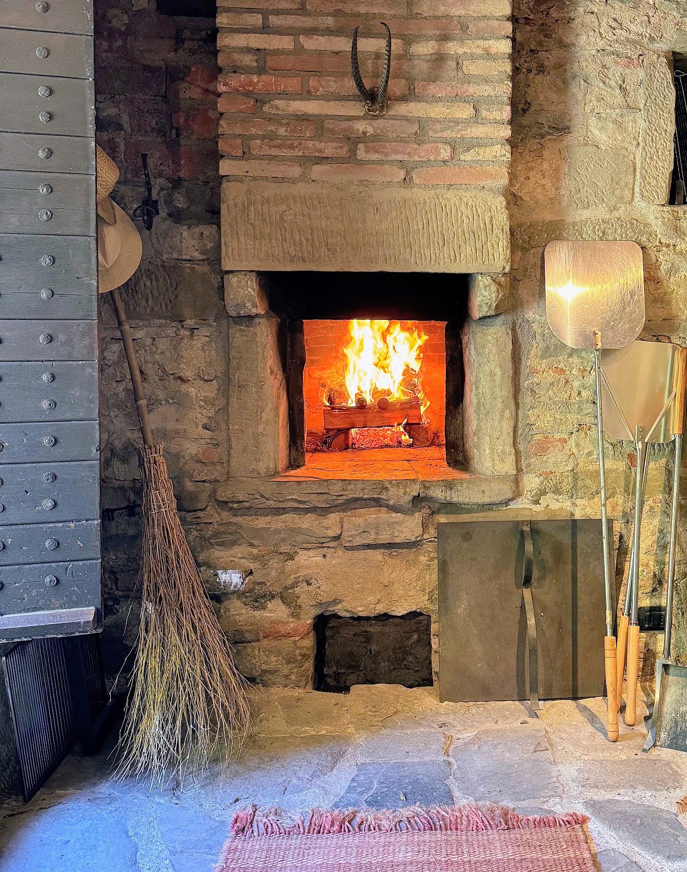 The lit mouth of the domed wood-fired oven with a fire burning inside, peels, a broom and tools leaning against the brick surround